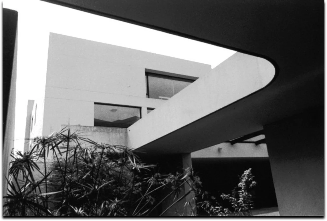 Vista en blanco y negro de un edificio moderno con ventanas rectangulares y un pasillo o pasarela en la parte superior, rodeado de plantas en primer plano.