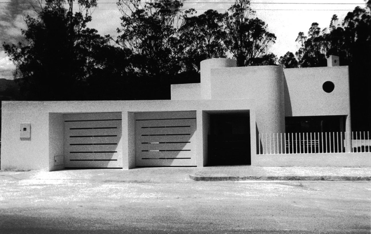 Casa moderna de color blanco con muros curvos y portones de garaje horizontales, ubicada en un área con árboles y cielo nublado.