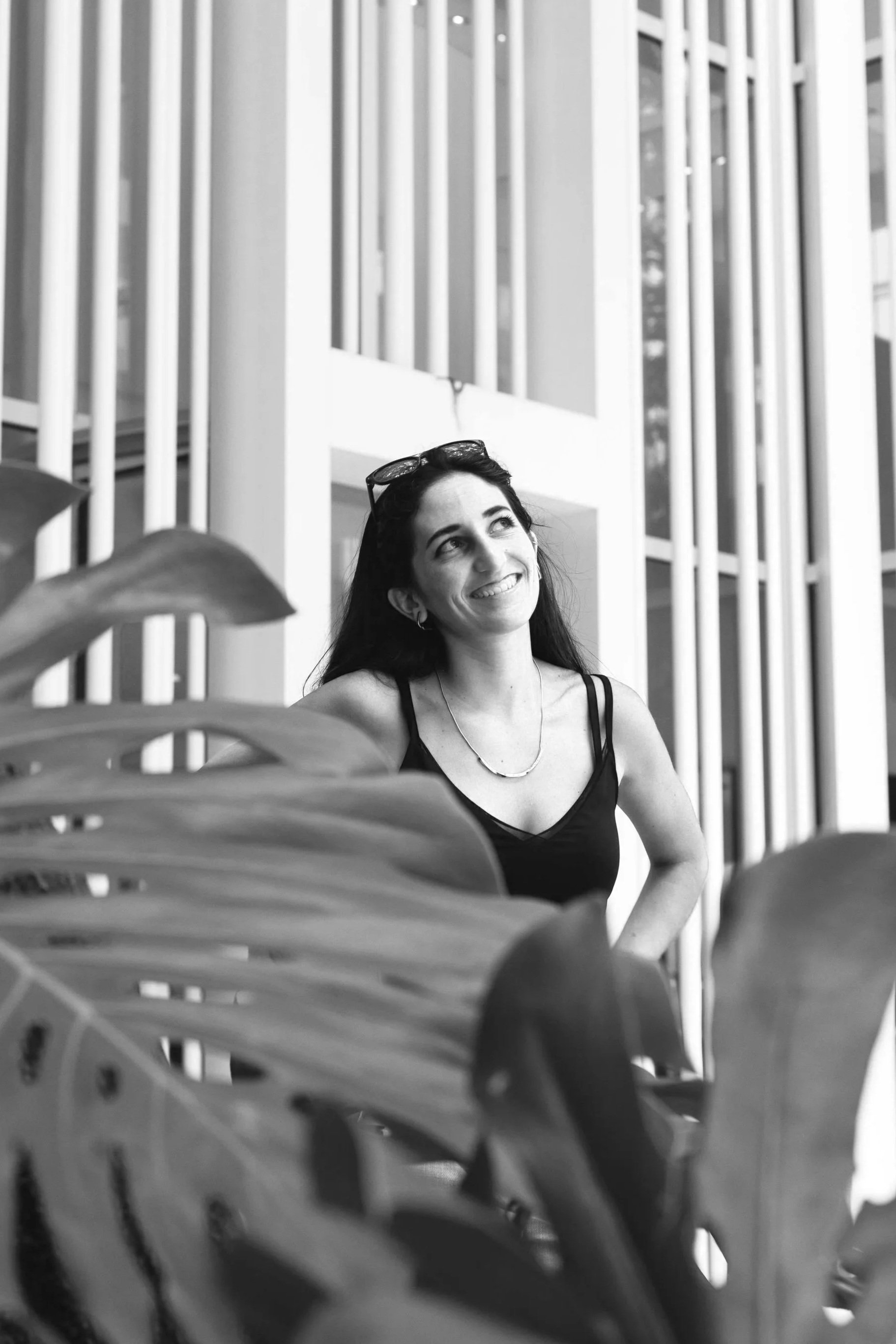 Mujer con cabello oscuro y gafas en la cabeza, sonriendo, en un espacio moderno con paredes de listones verticales, vista parcialmente bloqueada por plantas en primer plano.