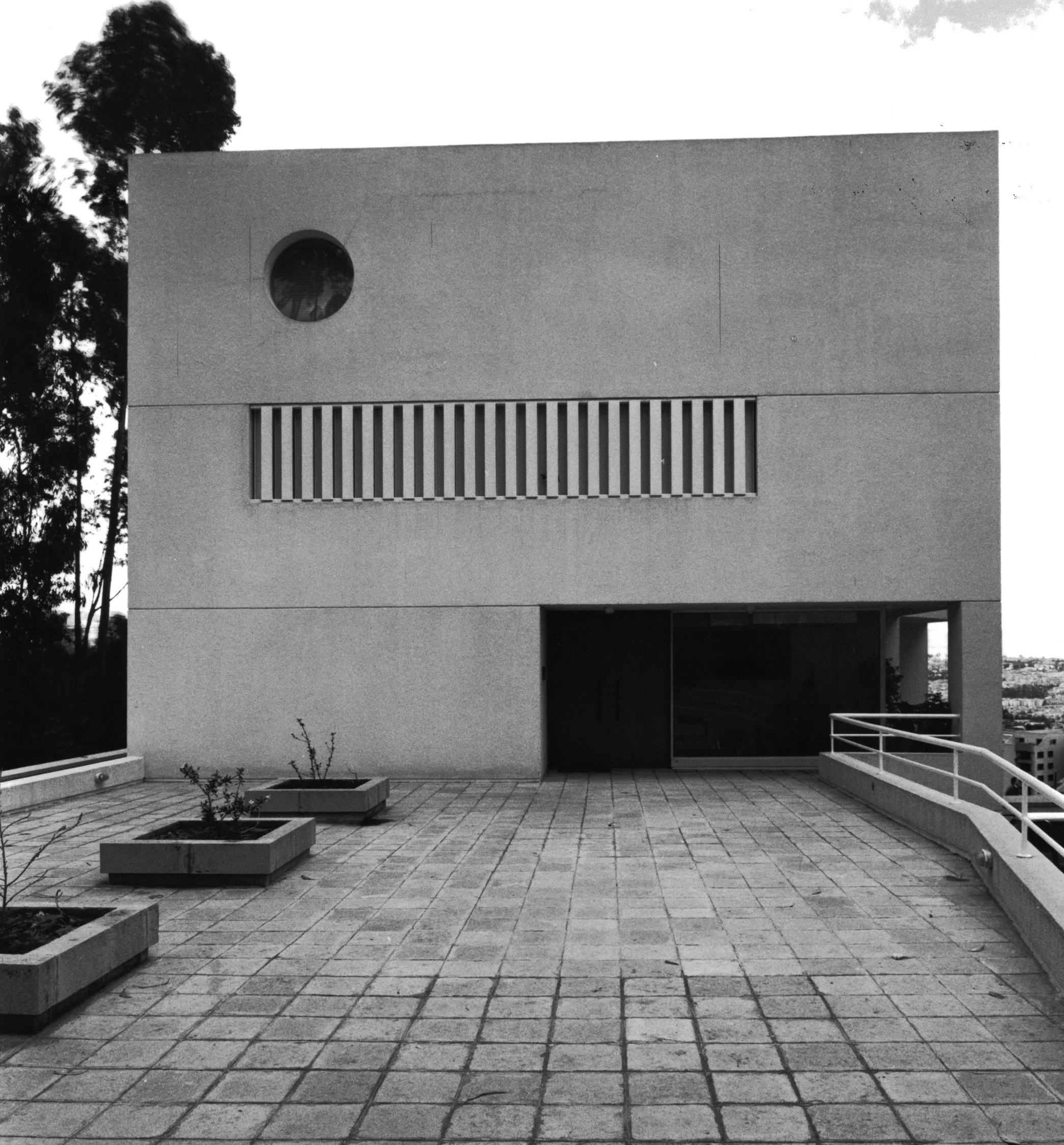 Edificio moderno de color blanco con ventanas rayadas y un círculo en la pared, en un patio con plantas y baldosas, rodeado de árboles
