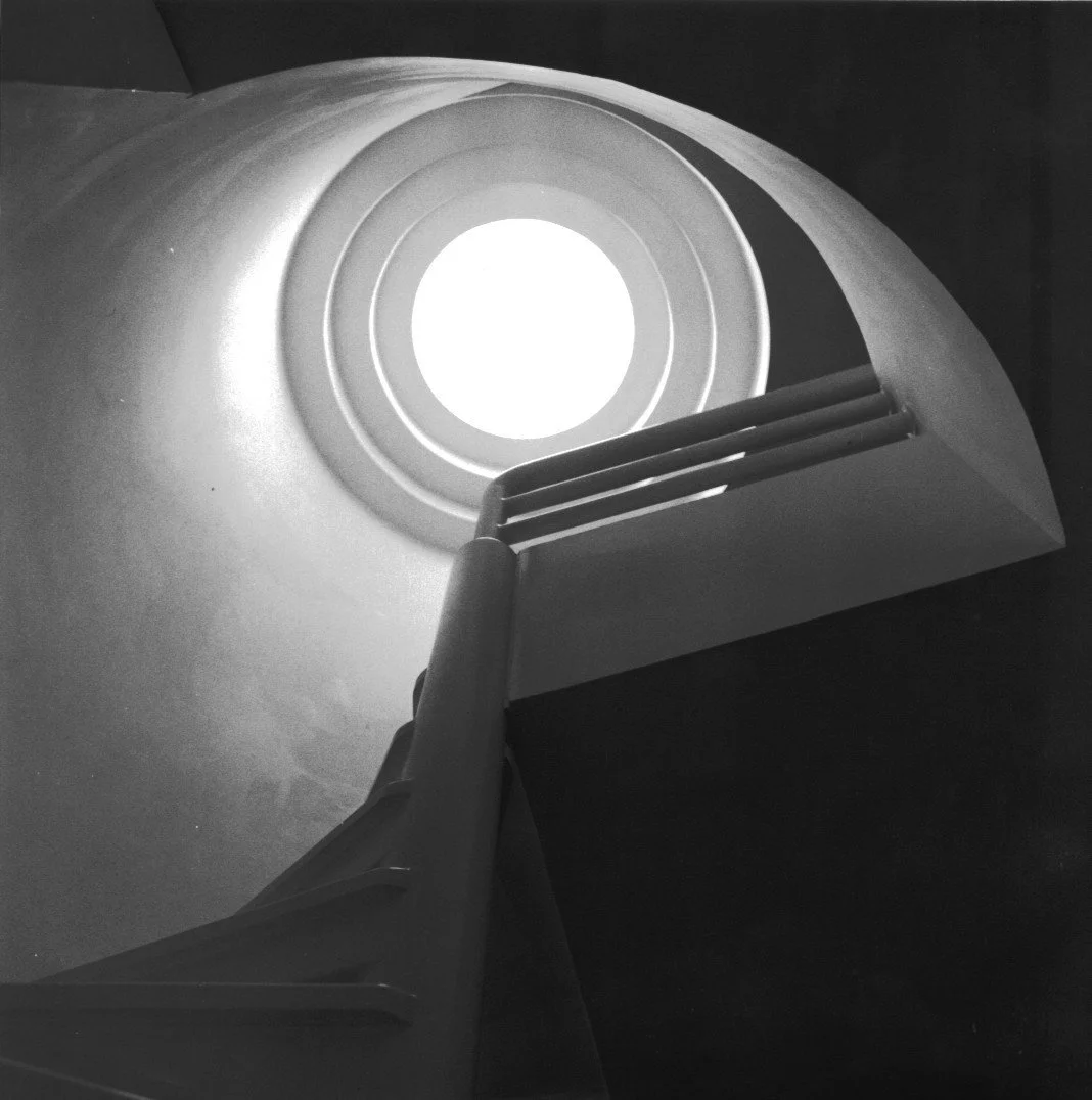 Vista desde abajo de una escalera que conduce a una abertura circular de luz en el techo, en blanco y negro.