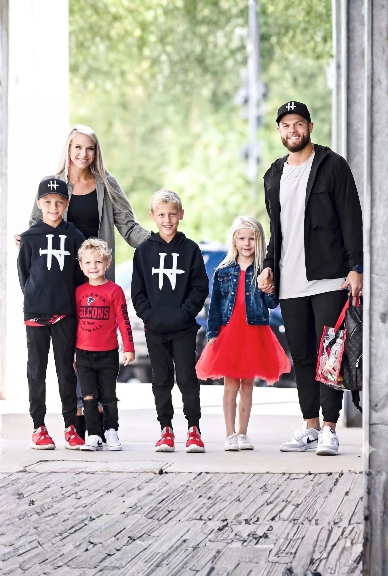 A group of six people, including three children and two adults, walking outdoors on a sidewalk with a blurred green background. The children are wearing casual clothes and sneakers, with two boys in black hoodies and a girl in a red dress with a denim jacket. The adults are smiling, with the woman in a gray jacket, and the man in a black jacket and cap. The group appears happy and engaged.