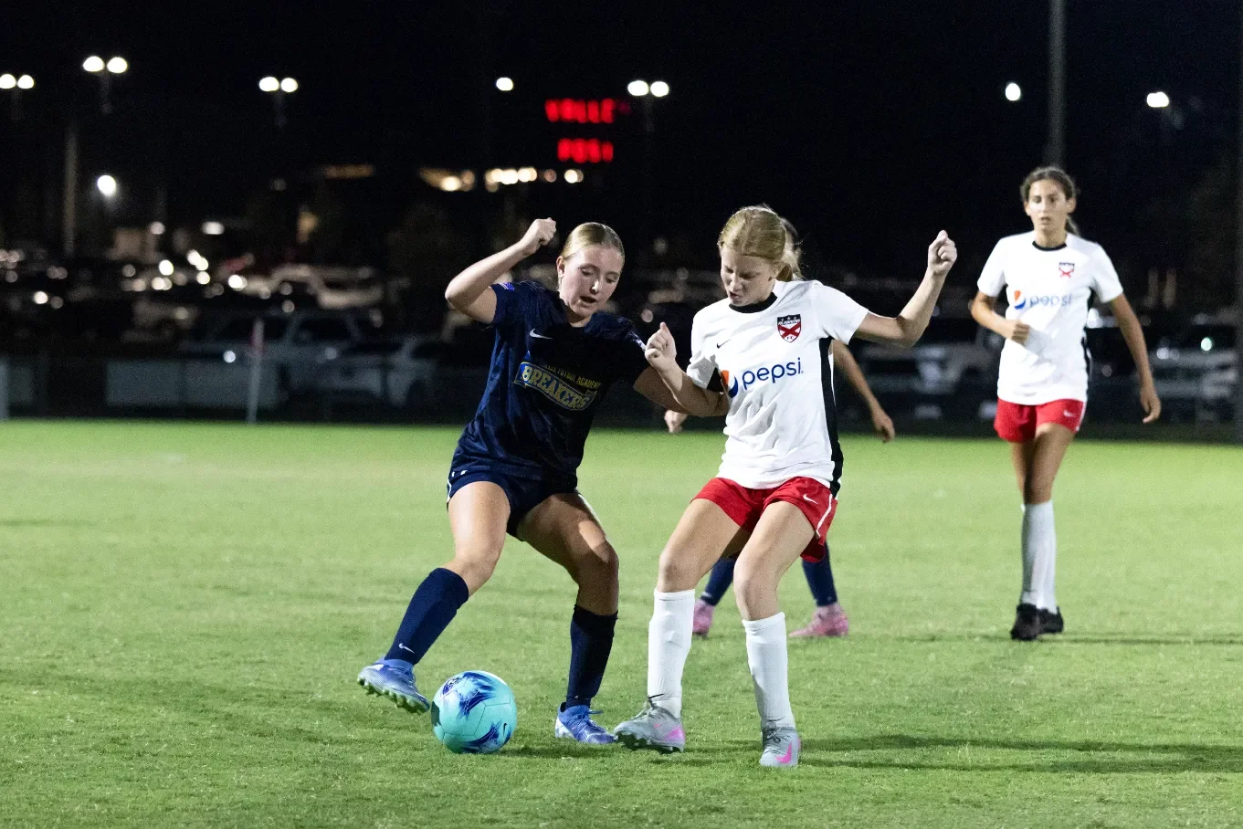 Two young female soccer players compete for the ball during a night game, with another player watching in the background, on a well-lit grass field.