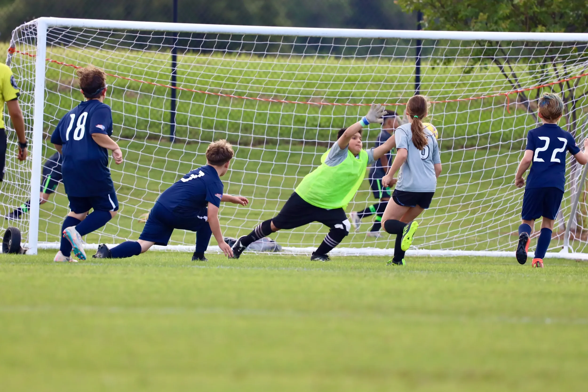 Children playing soccer in a grassy field, with a goalkeeper in a neon green jersey and white gloves hitting the ball into the goal as other kids watch and run nearby.