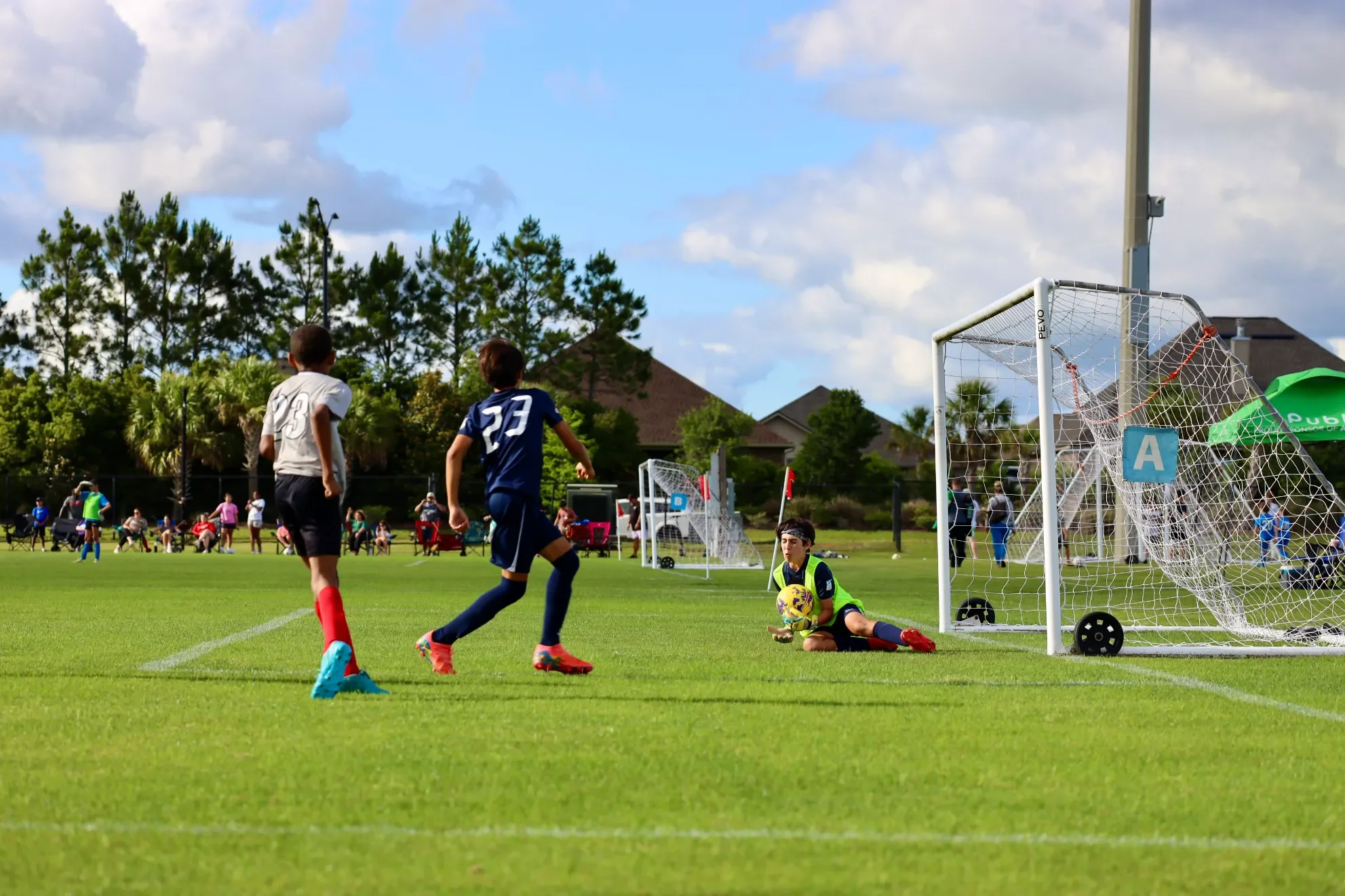 Children playing soccer on a field, with one goalkeeper sitting on the ground holding a yellow soccer ball, while two other children approaching the goal, with spectators and houses in the background under a partly cloudy sky.