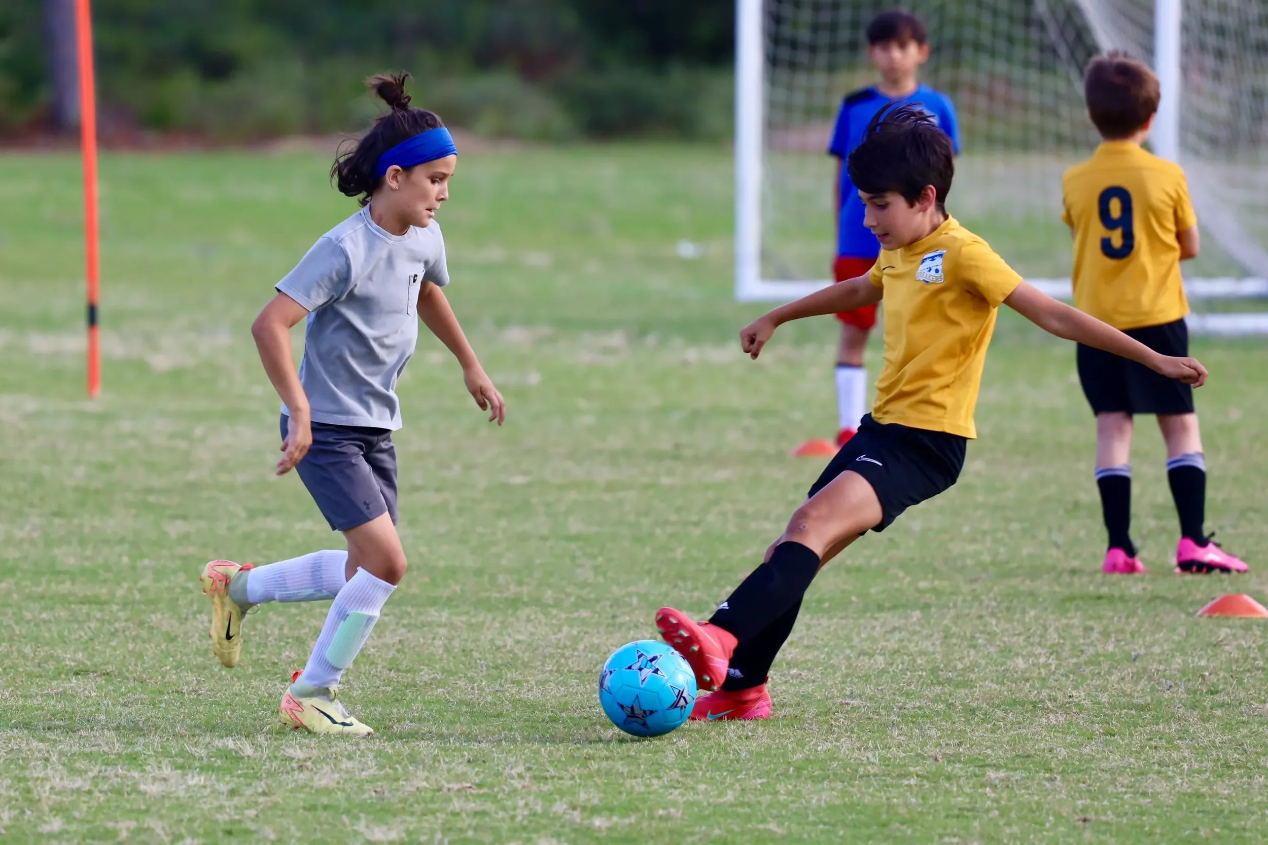 Children playing soccer on a grassy field with cones and goalposts.