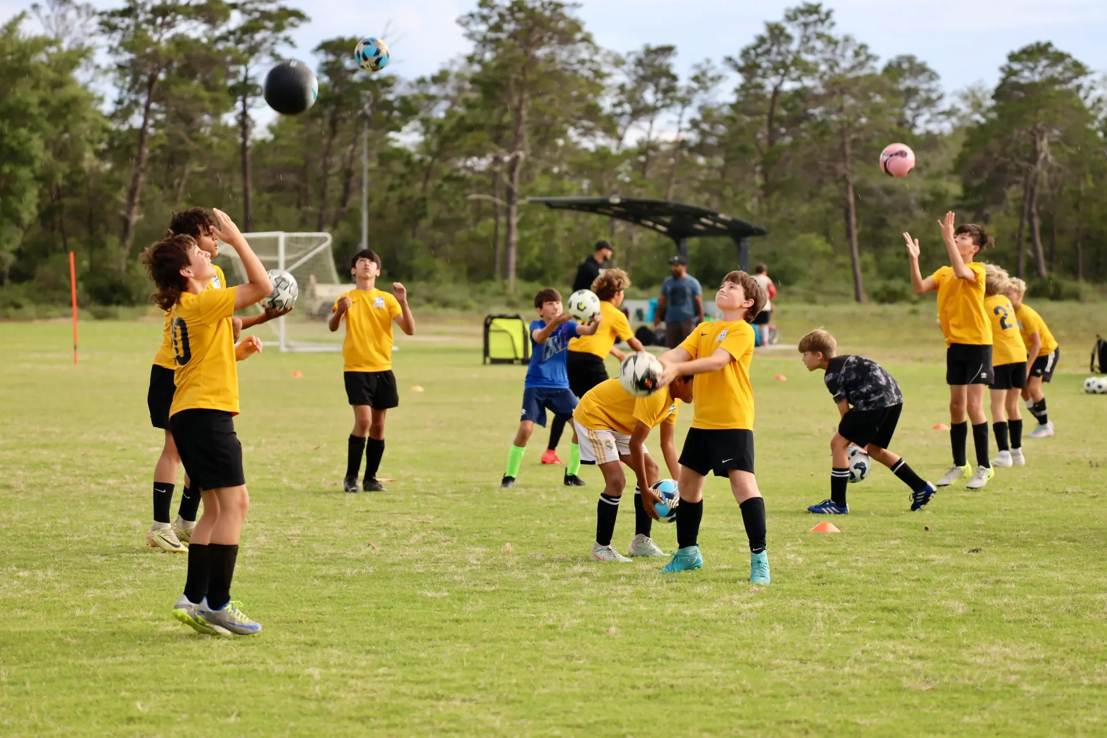 Children in yellow and black soccer uniforms practicing with balls on a field, some children throwing or catching soccer balls while a few are watching or preparing for their turn, with trees and a sports goal in the background.