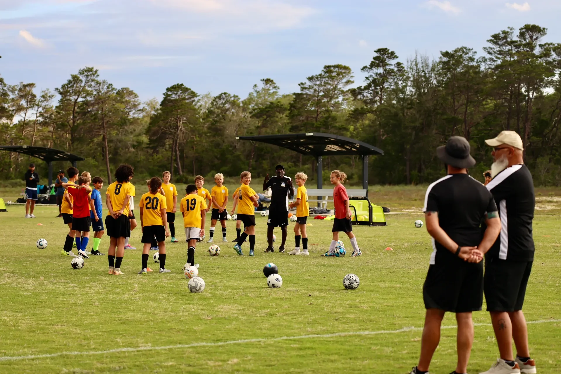 Youth soccer team practicing on a grassy field with coaches and instructors, in front of a backdrop of trees.