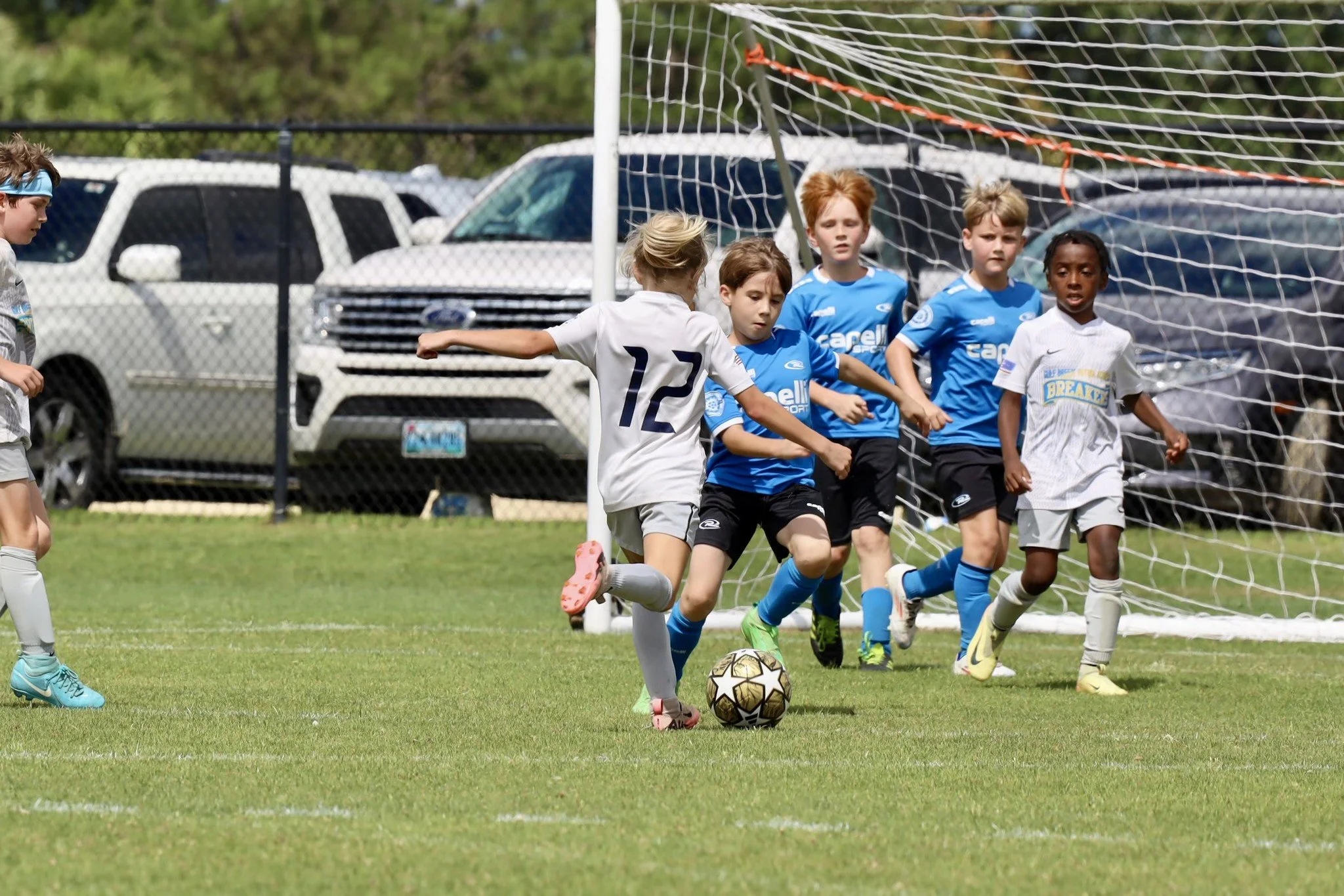 Young children playing soccer on a grass field with a soccer ball near a goal, surrounded by park cars and a chain-link fence.