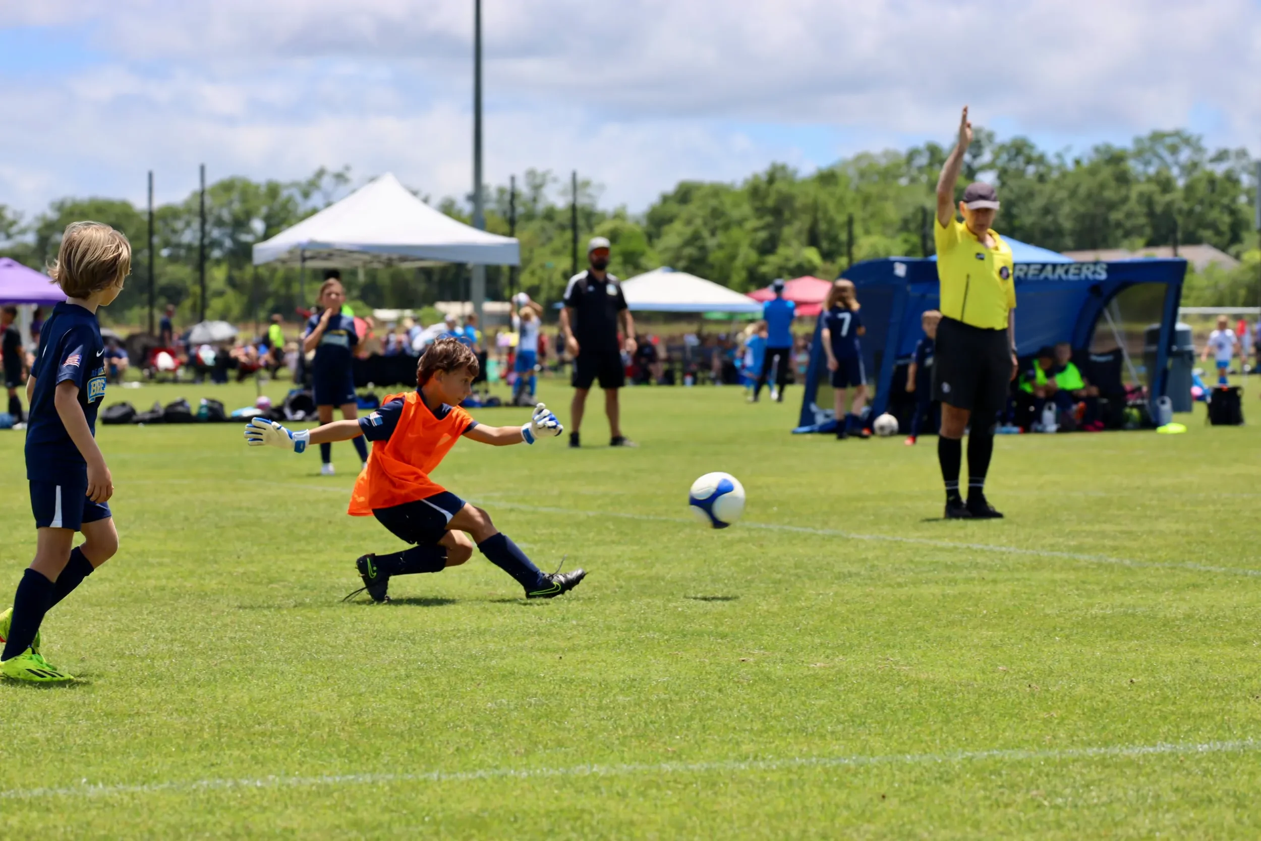 Children playing soccer on a grassy field during a sunny day, with tents and spectators in the background.