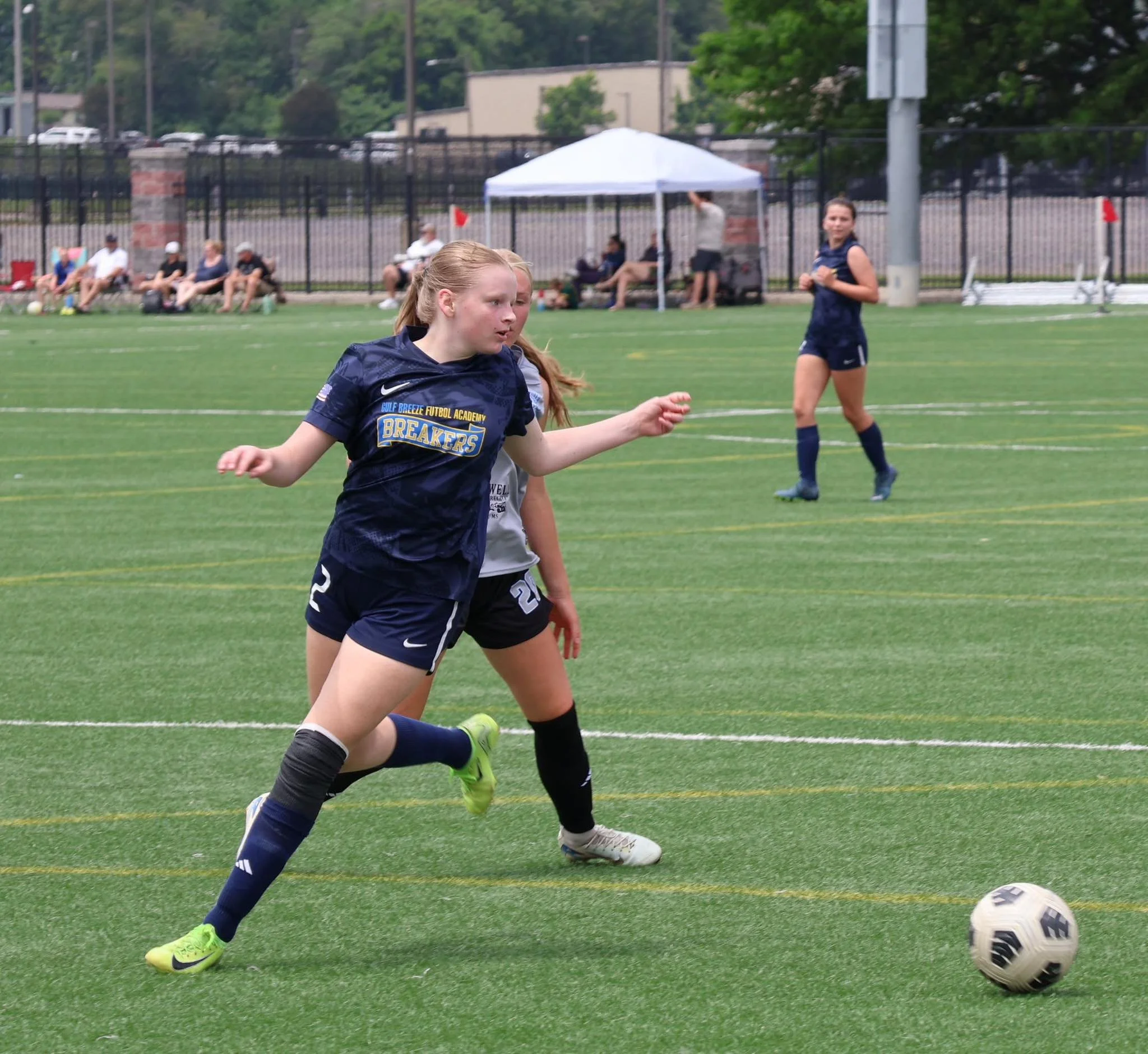 A young female soccer player with blonde hair in a ponytail, wearing a navy blue jersey with the words "Gulf Breeze Futbol Academy" and "Breakers" logo, attempting to kick a soccer ball on a soccer field. In the background, other players and spectators are seated and standing near a fence, with a small white canopy tent nearby.