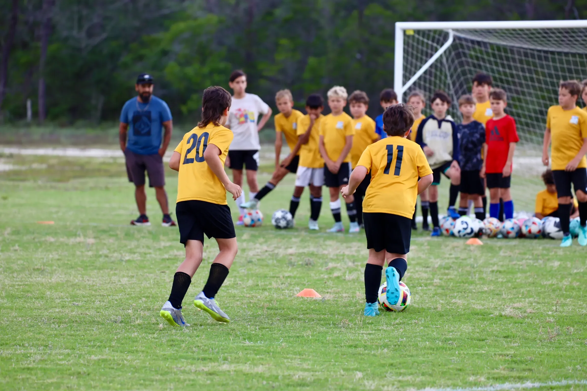 Children in yellow soccer jerseys practicing on a grassy field with a goalpost, soccer balls, and cones, supervised by coaches.