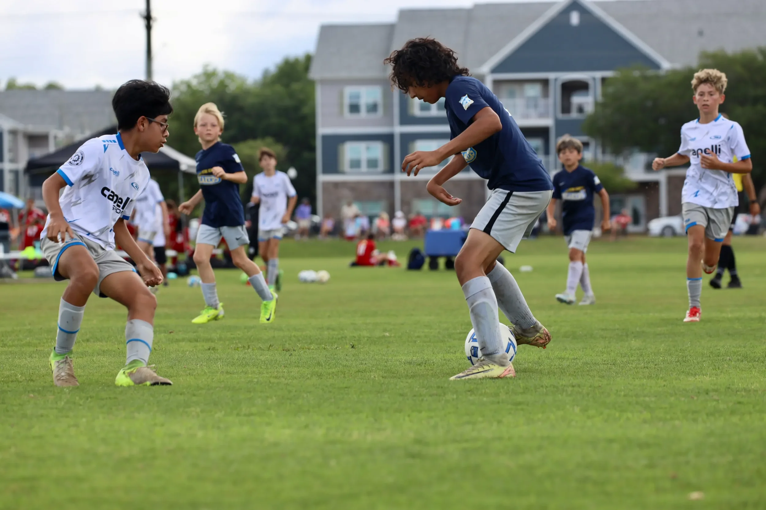 Children playing soccer on a grassy field with houses and trees in the background.