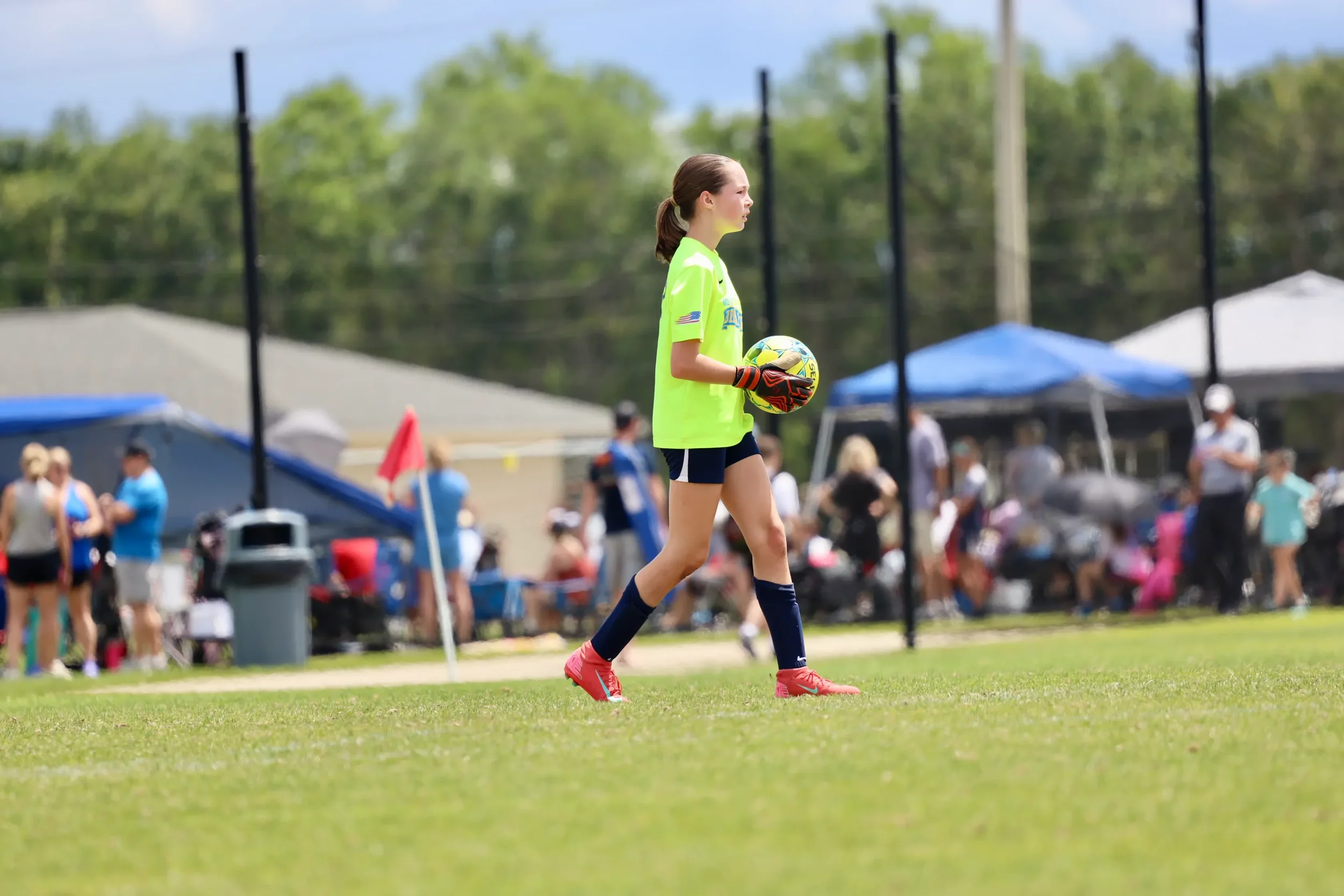 A young female soccer goalkeeper in bright yellow jersey, black shorts, pink cleats, and gloves holding a soccer ball on a grassy field during a game or practice, with spectators and tents in the background.
