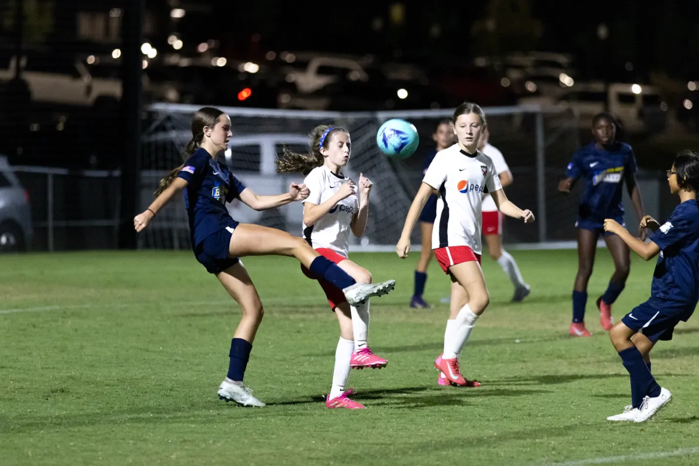 Girls playing soccer on a grass field at night, with some in blue uniforms and others in white and red uniforms, competing for the ball.
