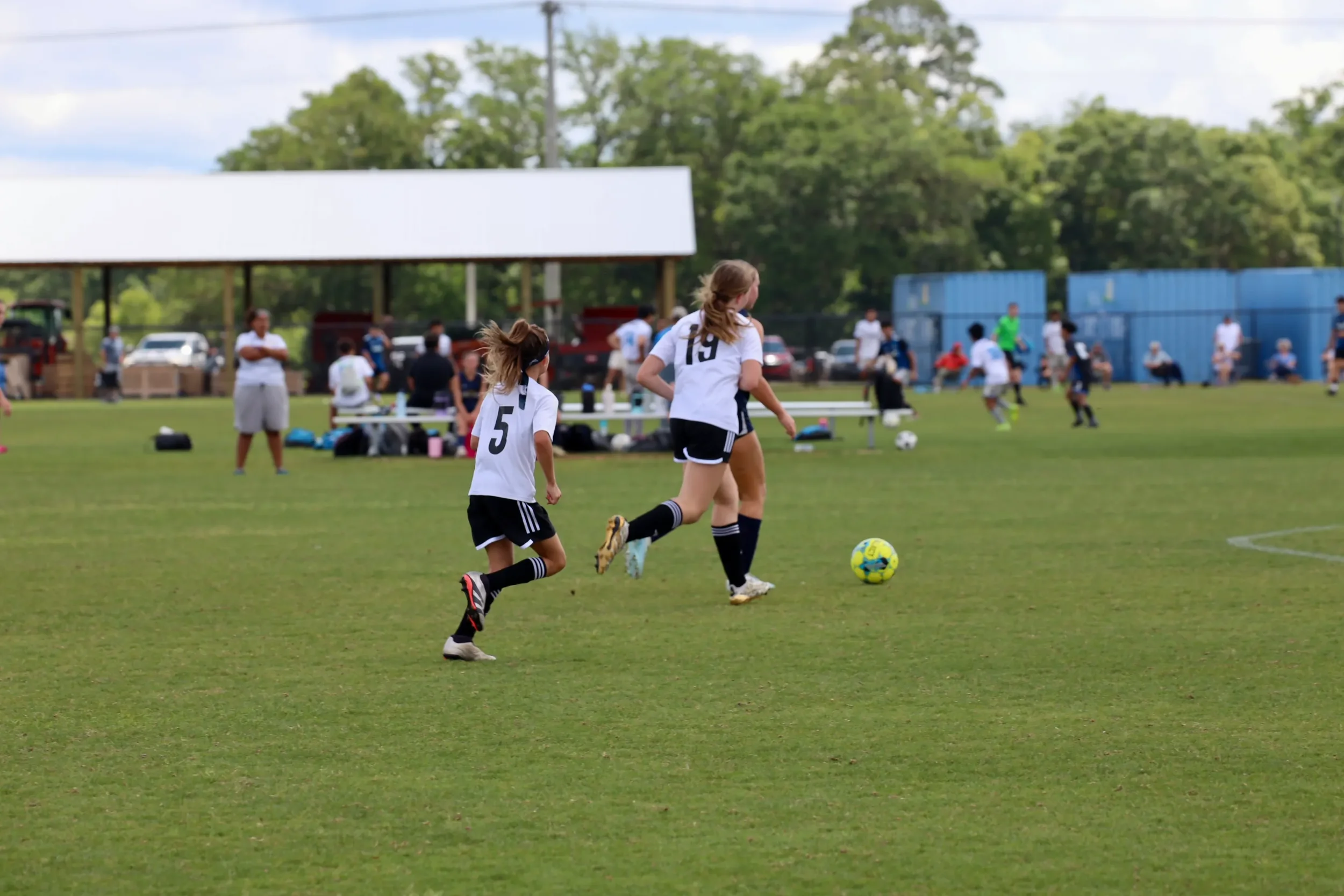 Two young girls playing soccer on a grassy field, with spectators and trees in the background.