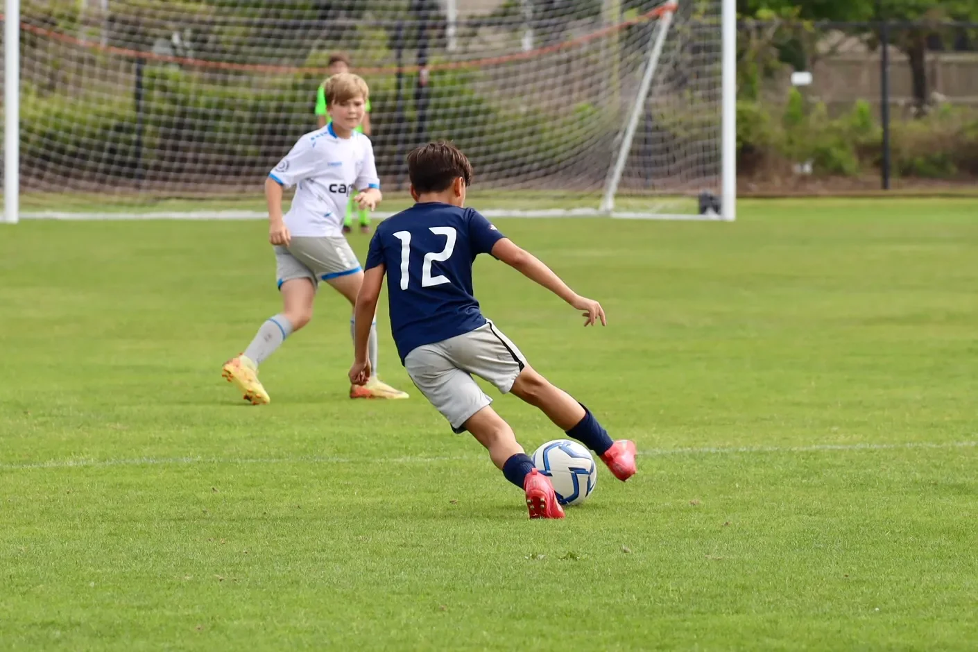 Young boy in a navy blue jersey and grey shorts kicking a soccer ball on a field, with a girl in a white jersey and grey shorts approaching, and a soccer goal in the background.
