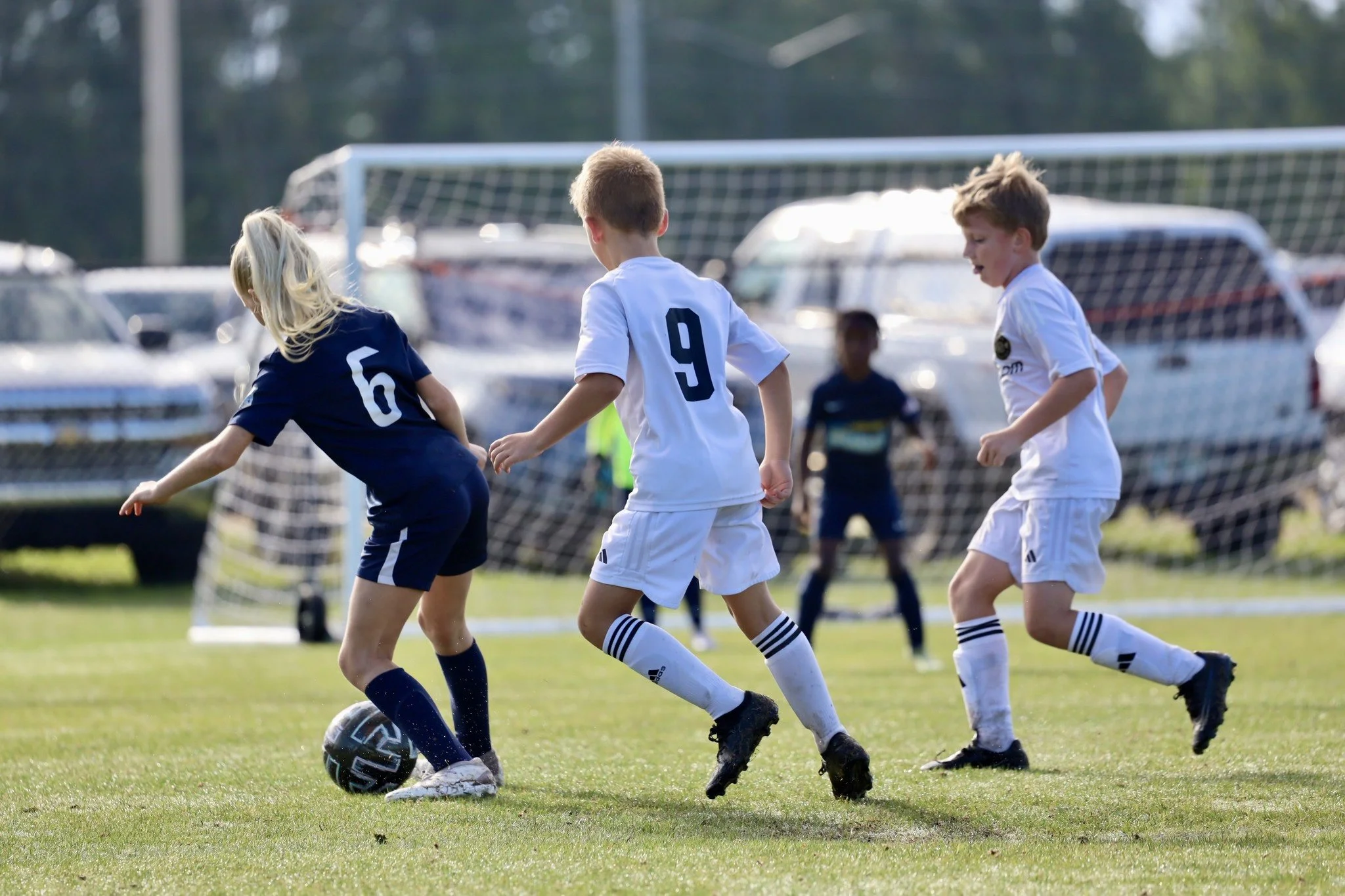 Children playing soccer on a field. One girl in a navy uniform with the number 6 is dribbling the ball, while boys in white uniforms are attempting to block her. A goalpost and parked cars are visible in the background.