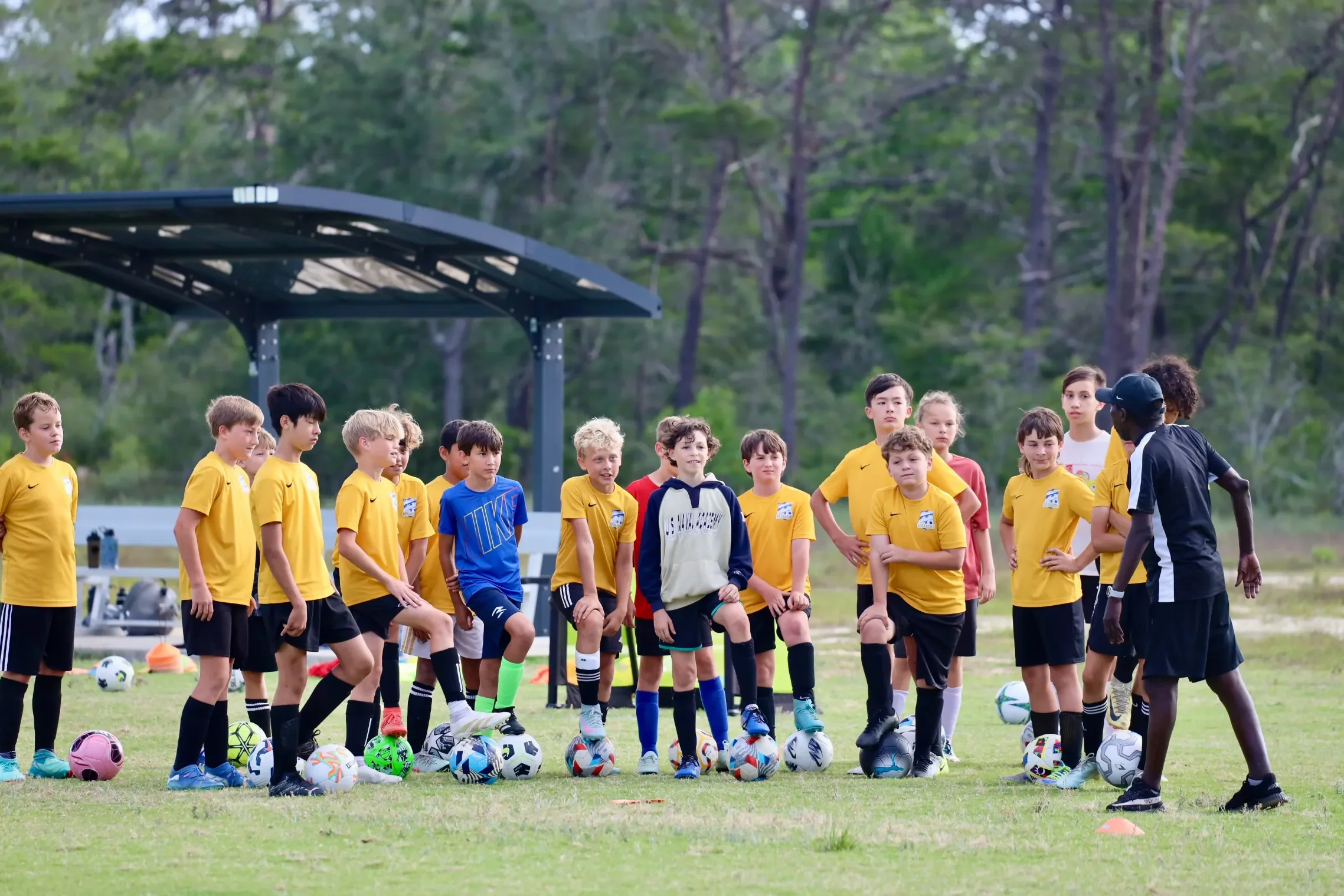 A group of young children, mostly boys in yellow and blue sports jerseys, standing on a soccer field during a practice or training session. A coach or instructor, dressed in black and white sportswear, is speaking to them. Soccer balls are lined up on the grass, and a shelter is visible in the background amidst trees.