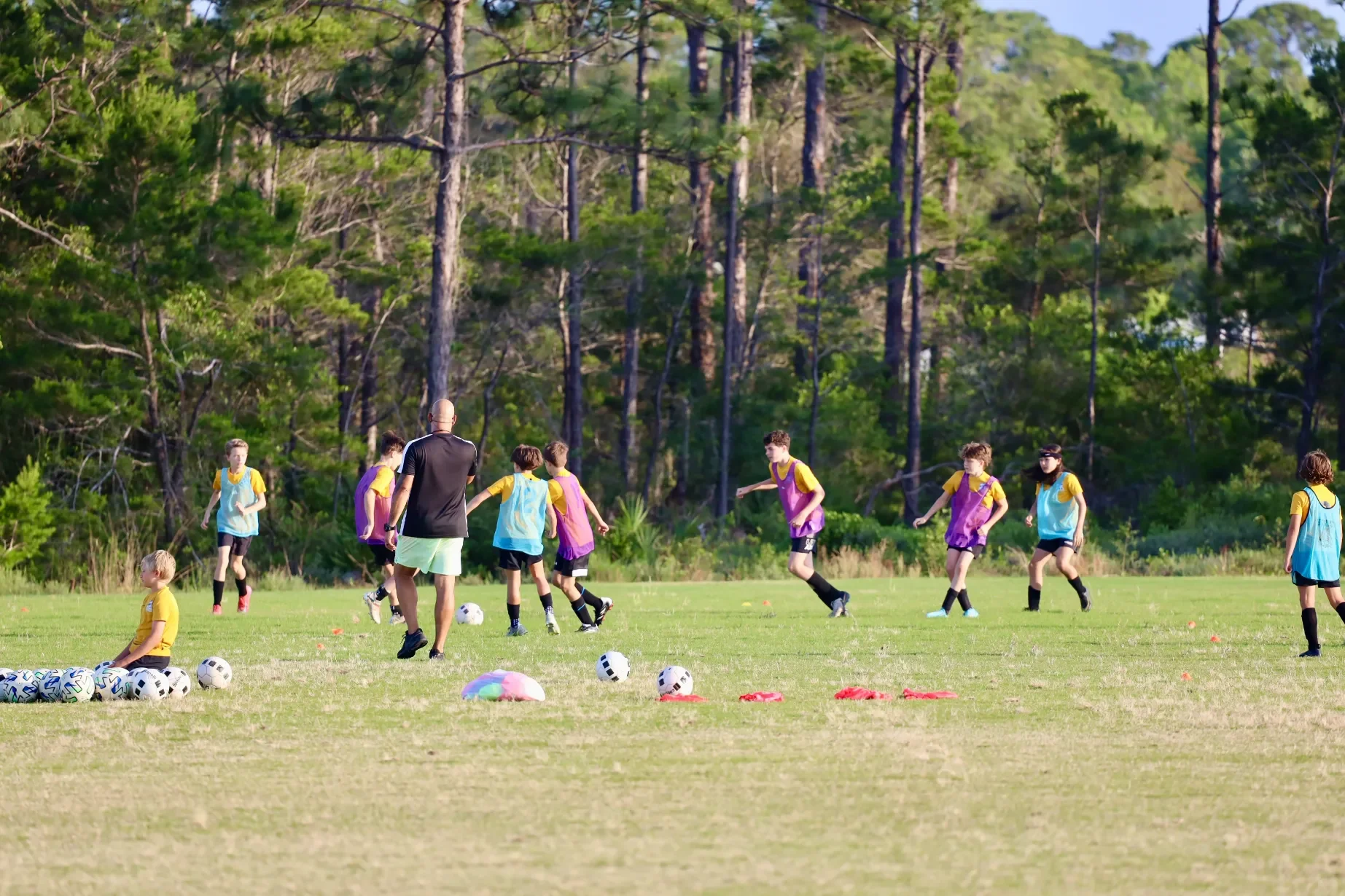 Children playing soccer on a grassy field with a coach, with trees in the background.