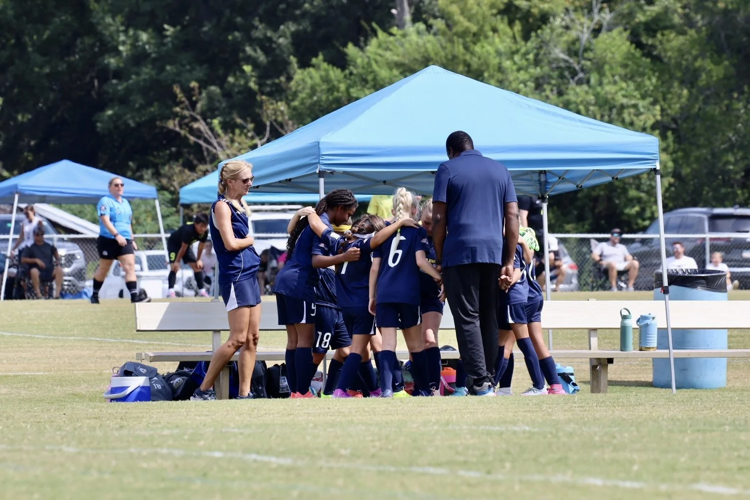 A youth soccer team huddle together on the field during a game, with a coach leading a prayer or discussion under a blue canopy. Other players and spectators are visible in the background.
