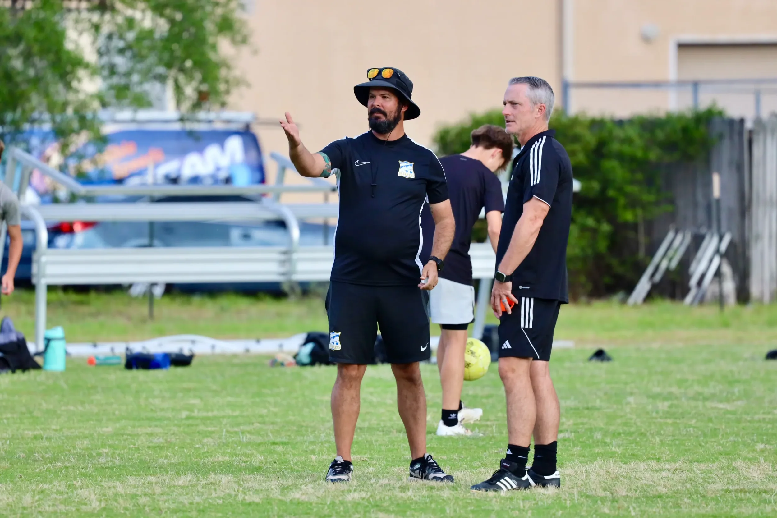 Two men standing on a soccer field talking, one gesturing with his hand. Both are wearing black sports shirts and shorts, and are surrounded by soccer equipment and a soccer ball. In the background, a person stands near a fence with trees and buildings beyond.