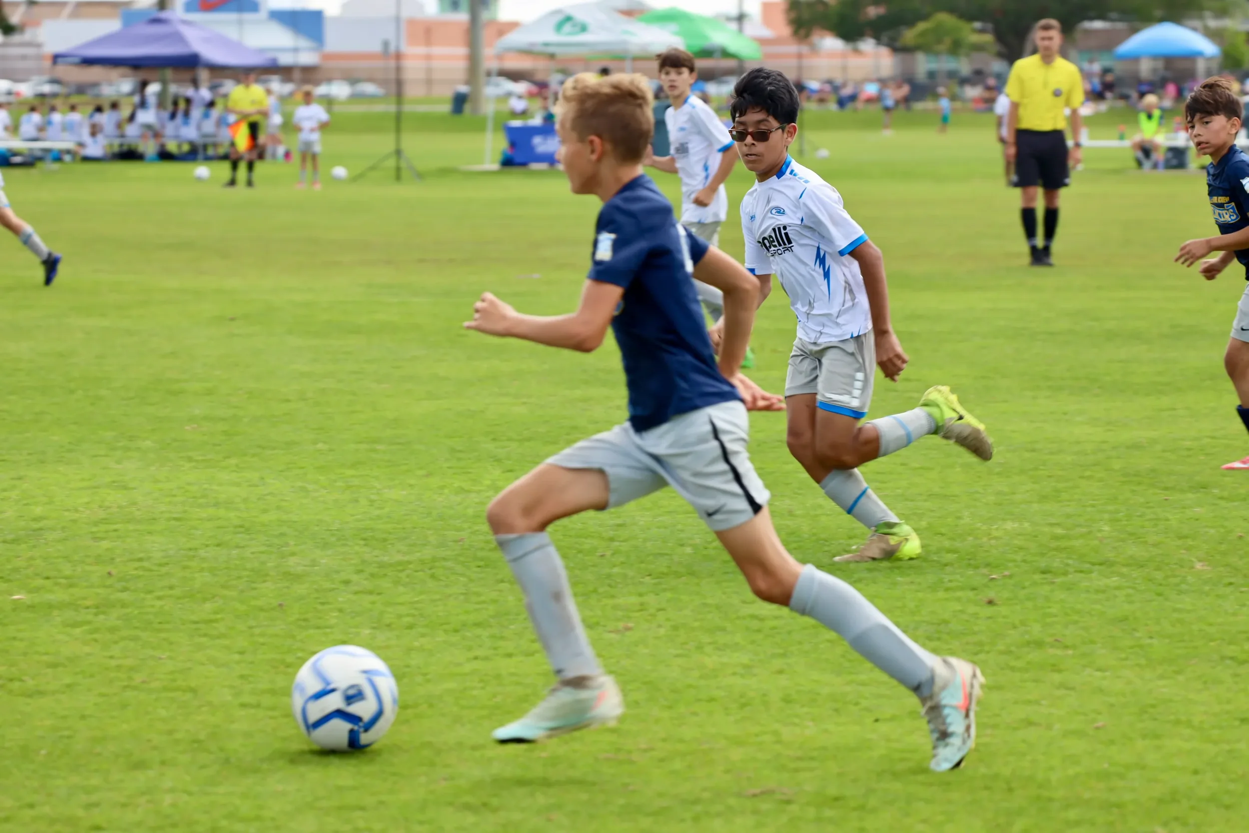 Youth soccer game with boys running after the ball on a grass field, surrounded by spectators and umbrellas.