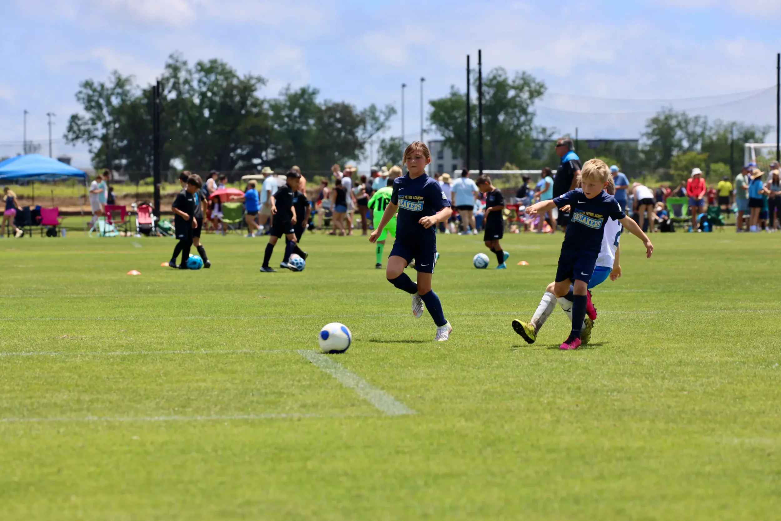 Children playing soccer on a grassy field with many spectators watching from the sidelines on a sunny day.