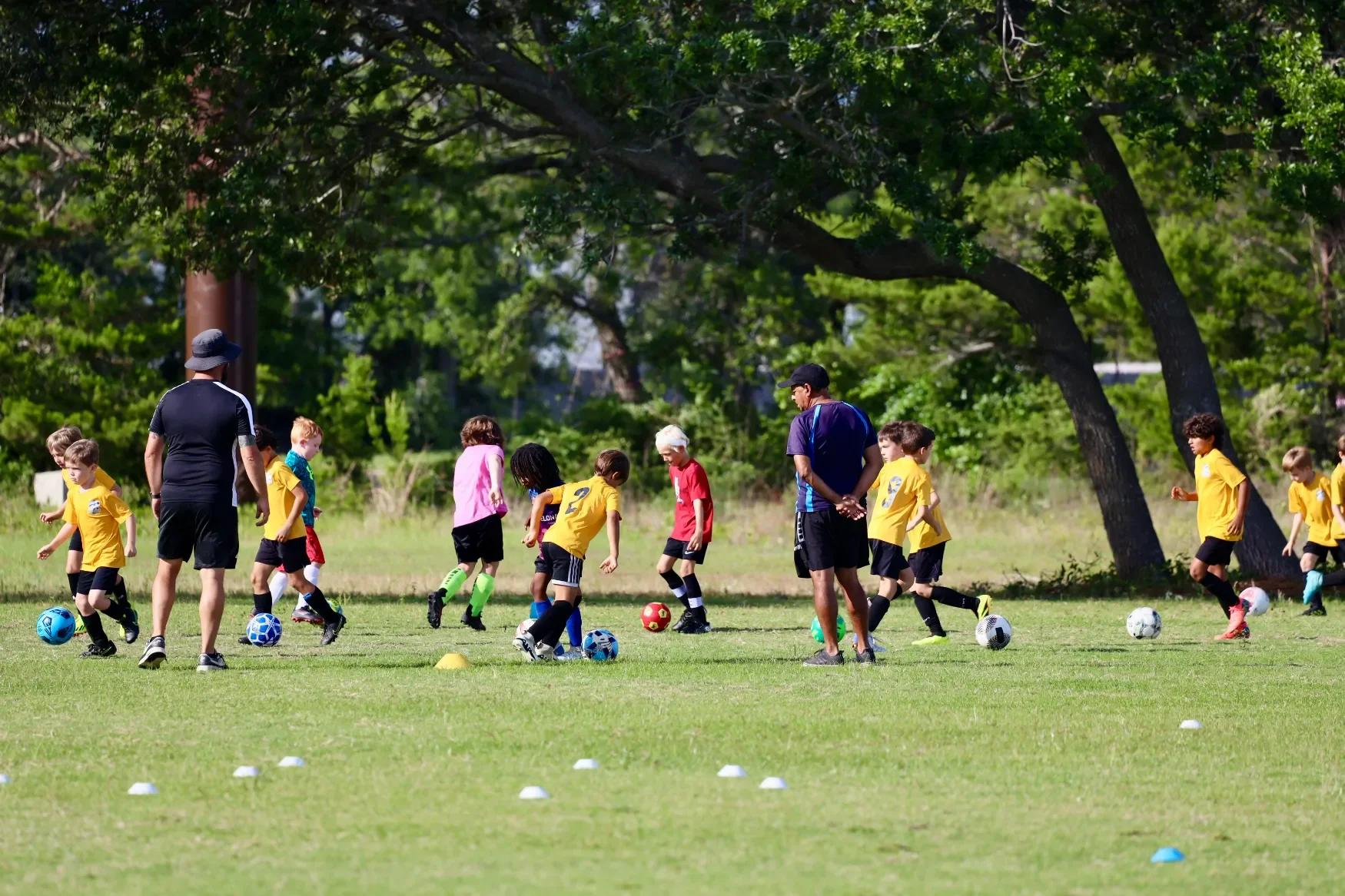 Children participating in a soccer practice on a grassy field under trees, with coaches present.
