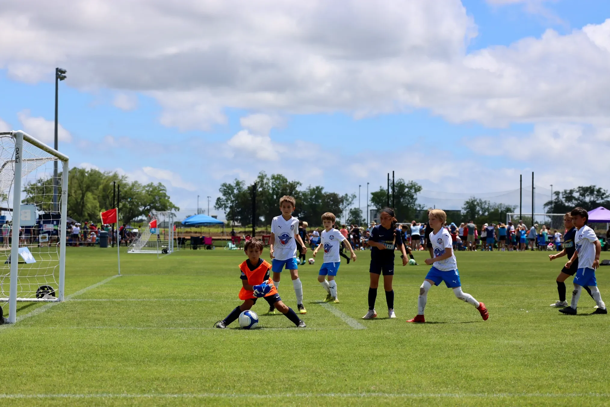 Children playing soccer during a youth soccer game on a sunny day, with spectators and tents in the background.