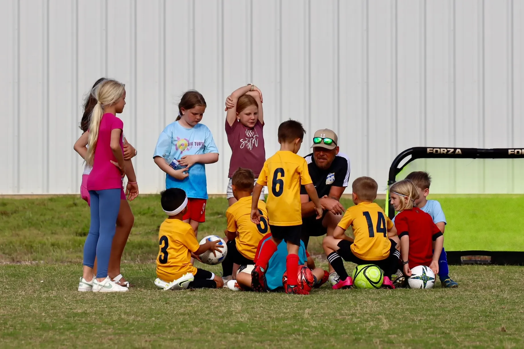 A group of young children on a soccer field gathered around their coach during a soccer practice or game. The children are wearing colorful sports jerseys and have soccer balls nearby.