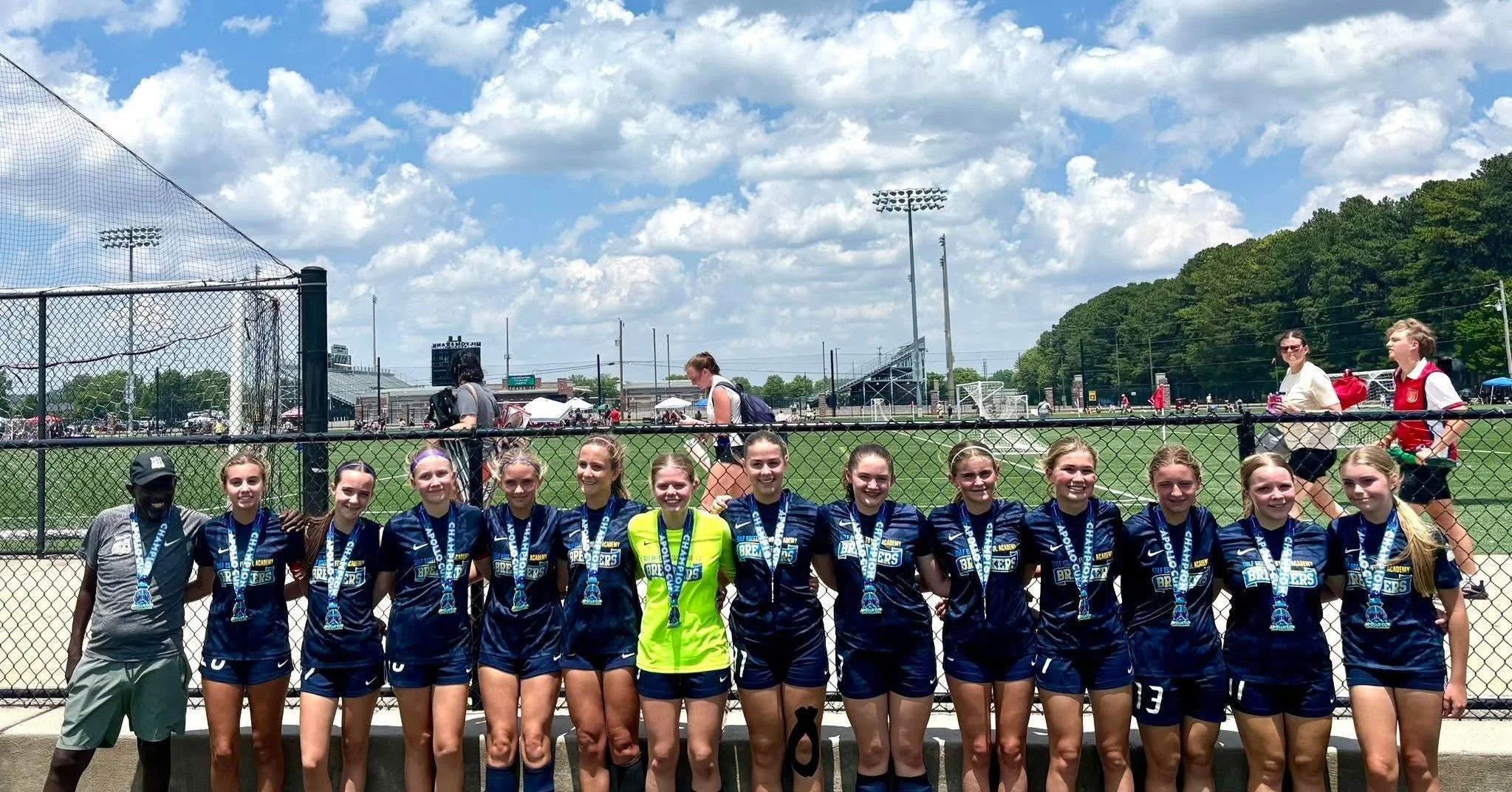 A girls' soccer team standing in front of a fence, wearing blue uniforms with medals around their necks, with a stadium and people in the background.