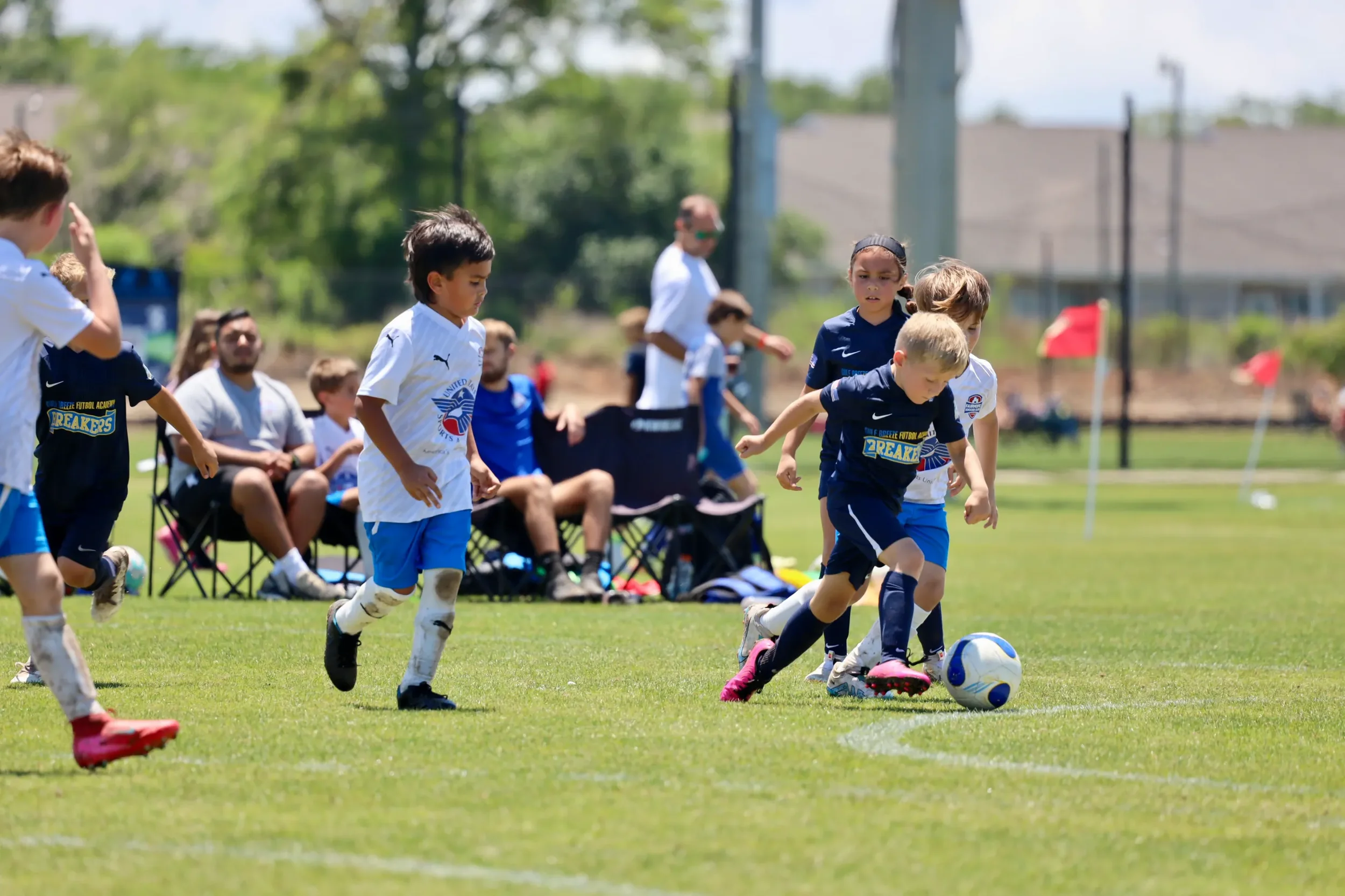 Children playing soccer on a grassy field during daytime, with spectators seated and standing in the background.