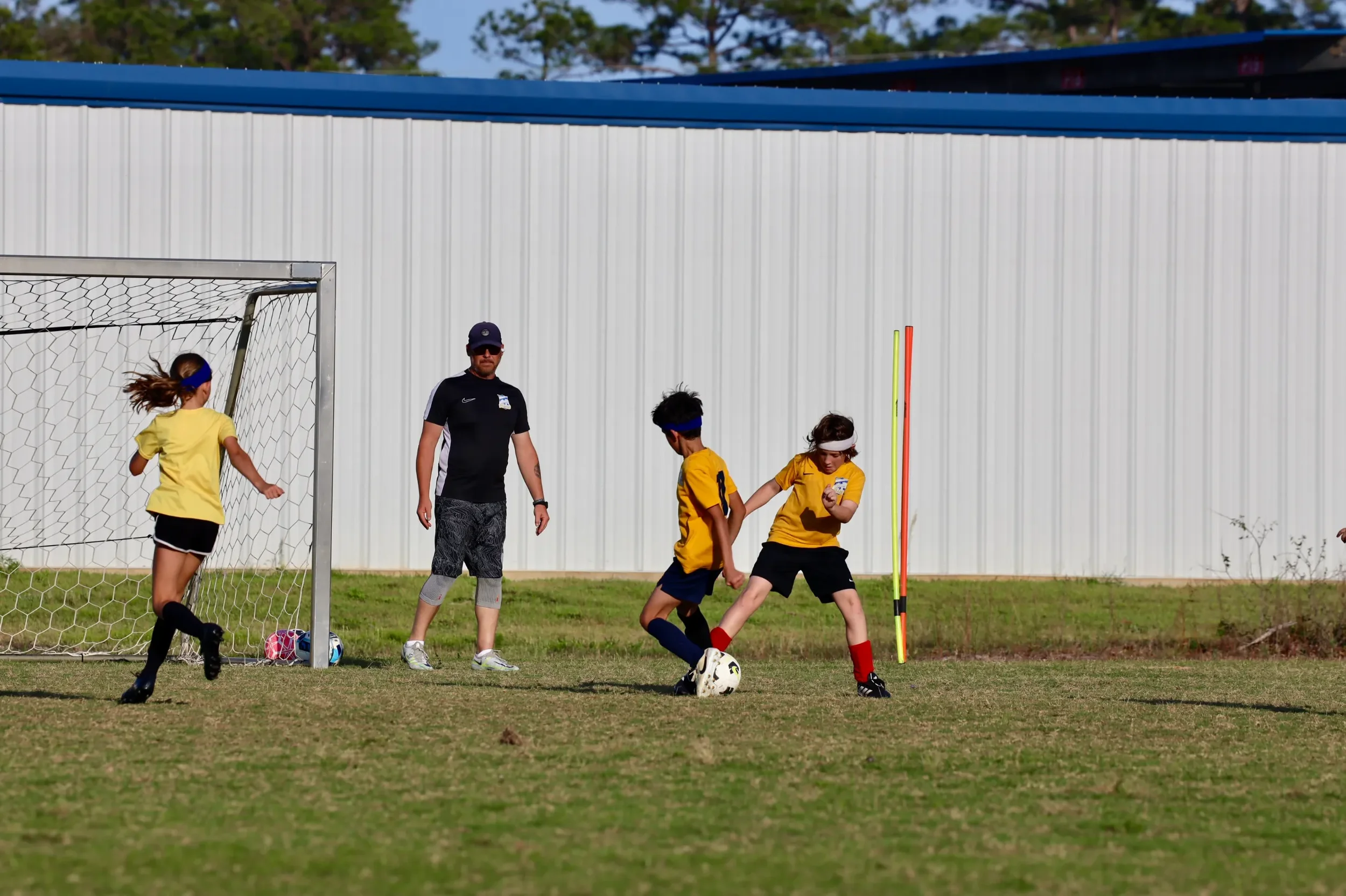 Children playing soccer on a field with a coach overseeing, near a goalpost, with a large white industrial building in the background.