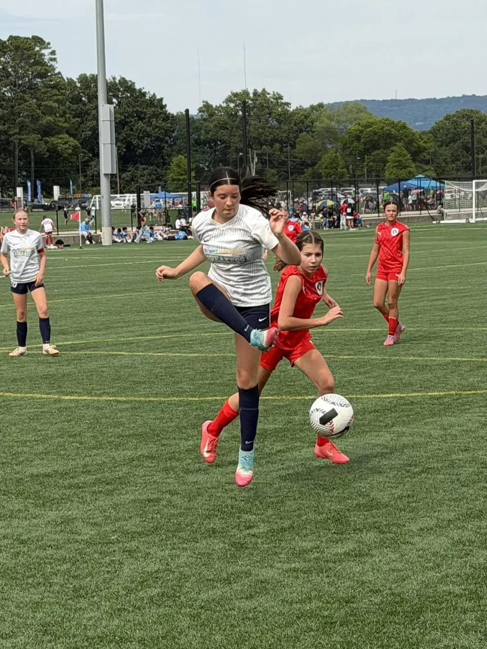 Girls playing soccer on a field, one girl in a white jersey and navy shorts jumping over a girl in a red jersey and shorts to kick a soccer ball.