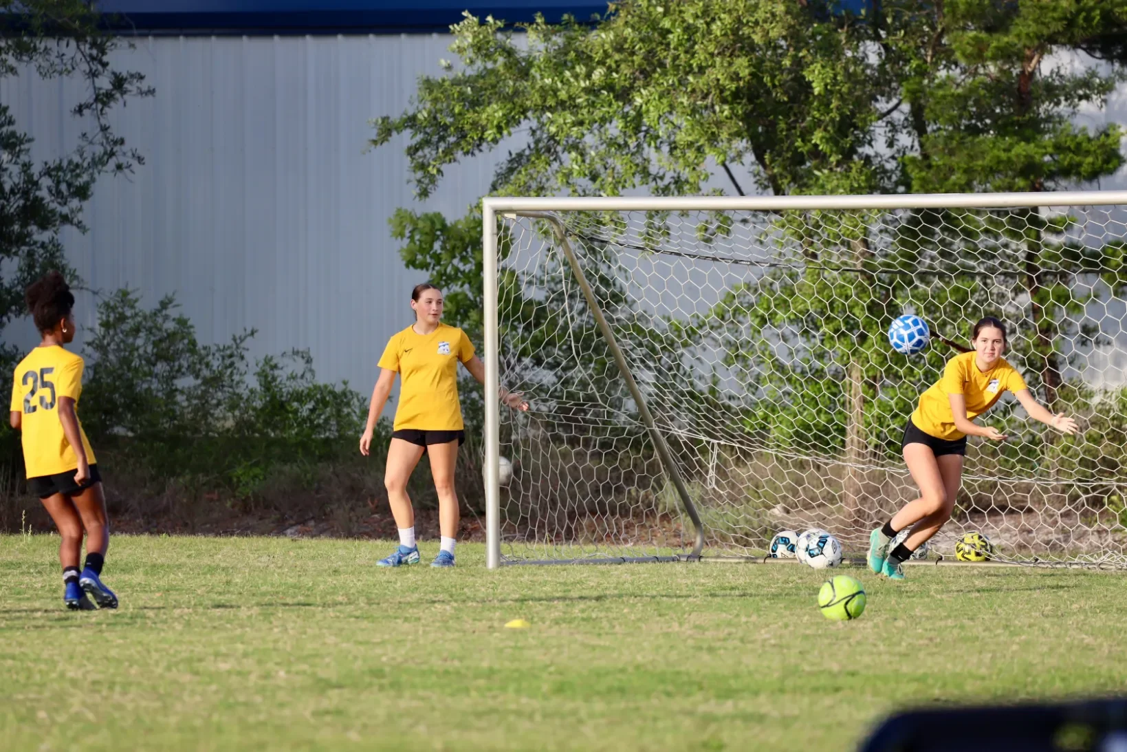 Young women playing soccer, with one about to kick a ball into the goal, while others stand nearby on a grassy field, wearing yellow jerseys and athletic shorts, with soccer balls and equipment scattered around.