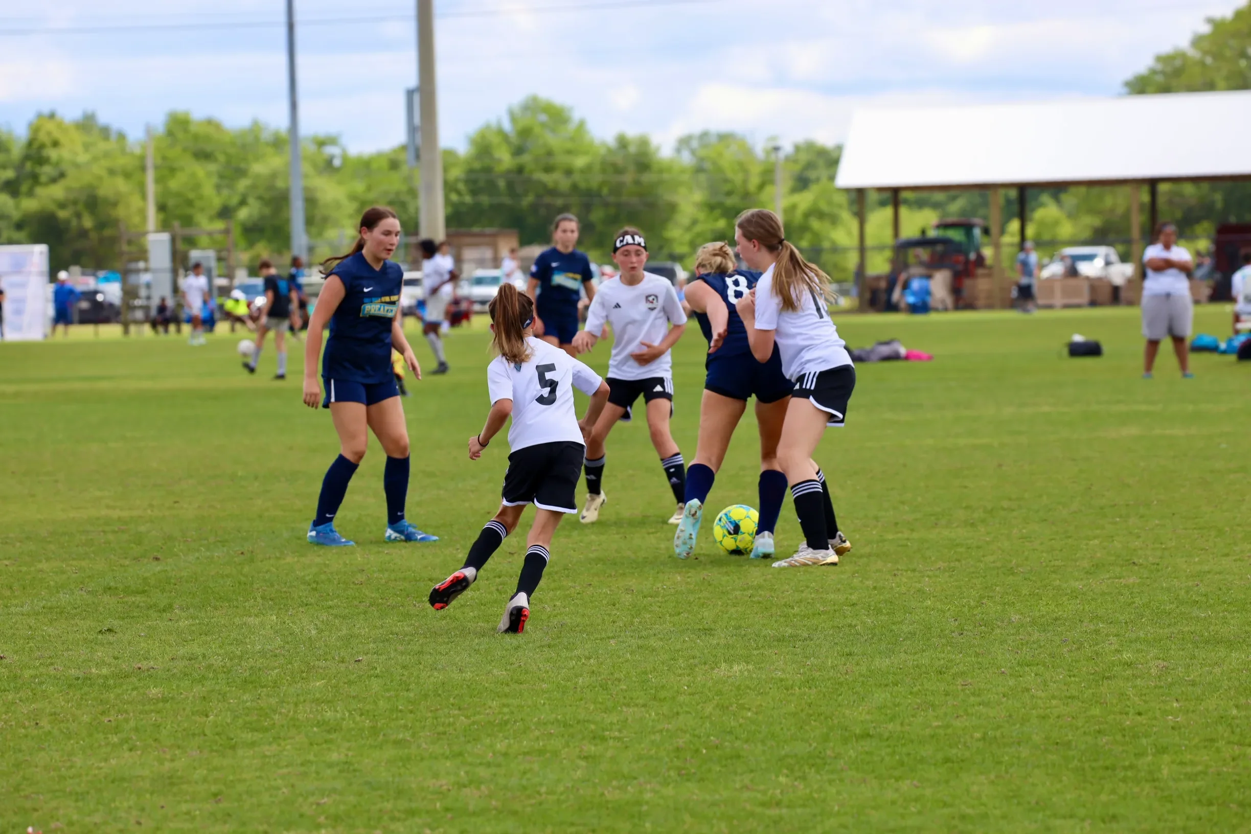 Young girls and women playing soccer on a grassy field during daytime, with trees and a pavilion in the background.