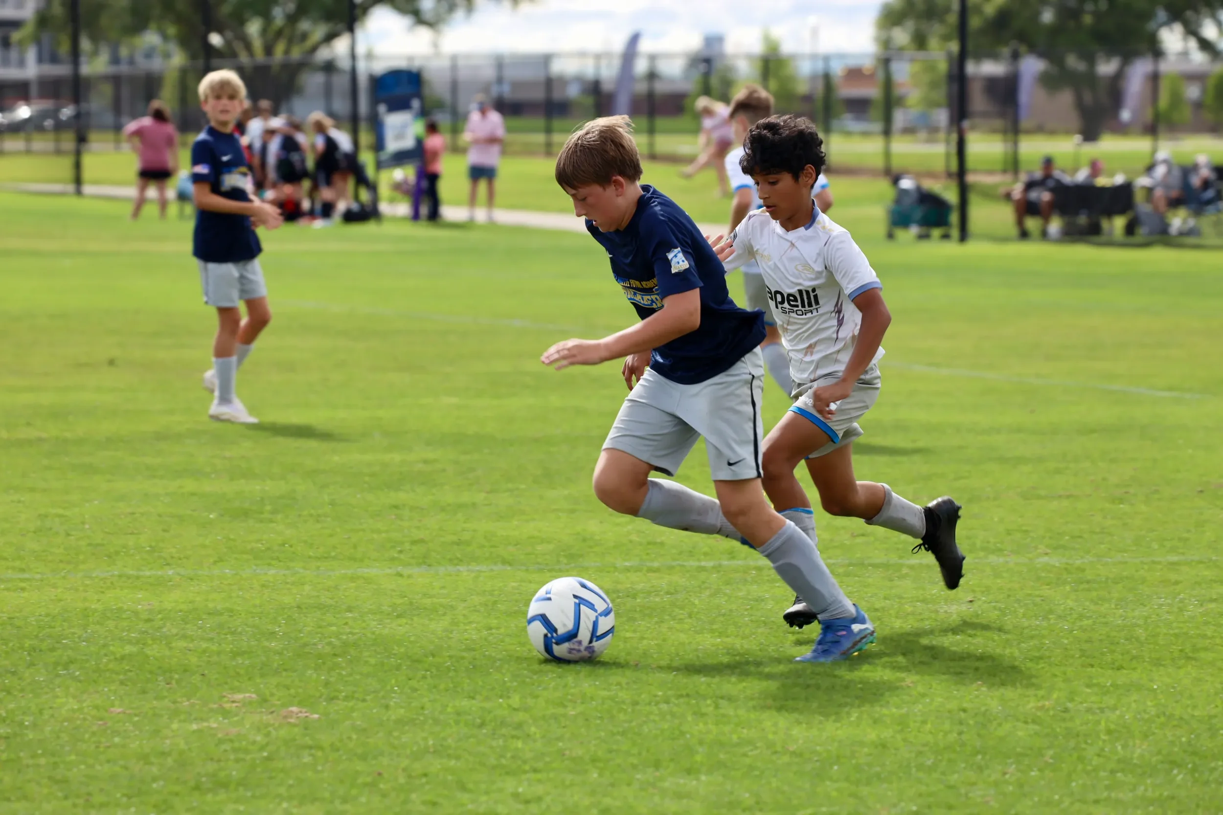Two young boys playing soccer on a green field with other children and spectators in the background.