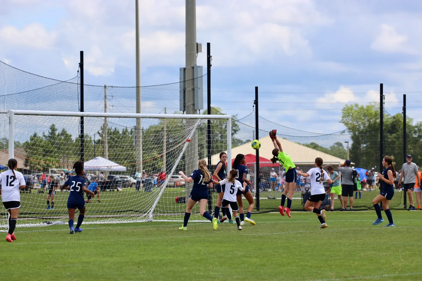 Girls' soccer game with players in navy and white jerseys, one goalie in bright green reaching for a ball, on a grassy field with crowd and tents in background.