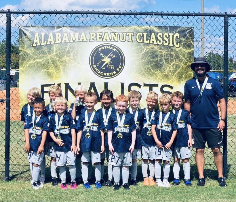 A youth soccer team with medals standing in front of a banner that reads 'Alabama Peanut Classic Finals' at a sports field. The team is wearing matching uniforms and an adult coach is standing to the right.
