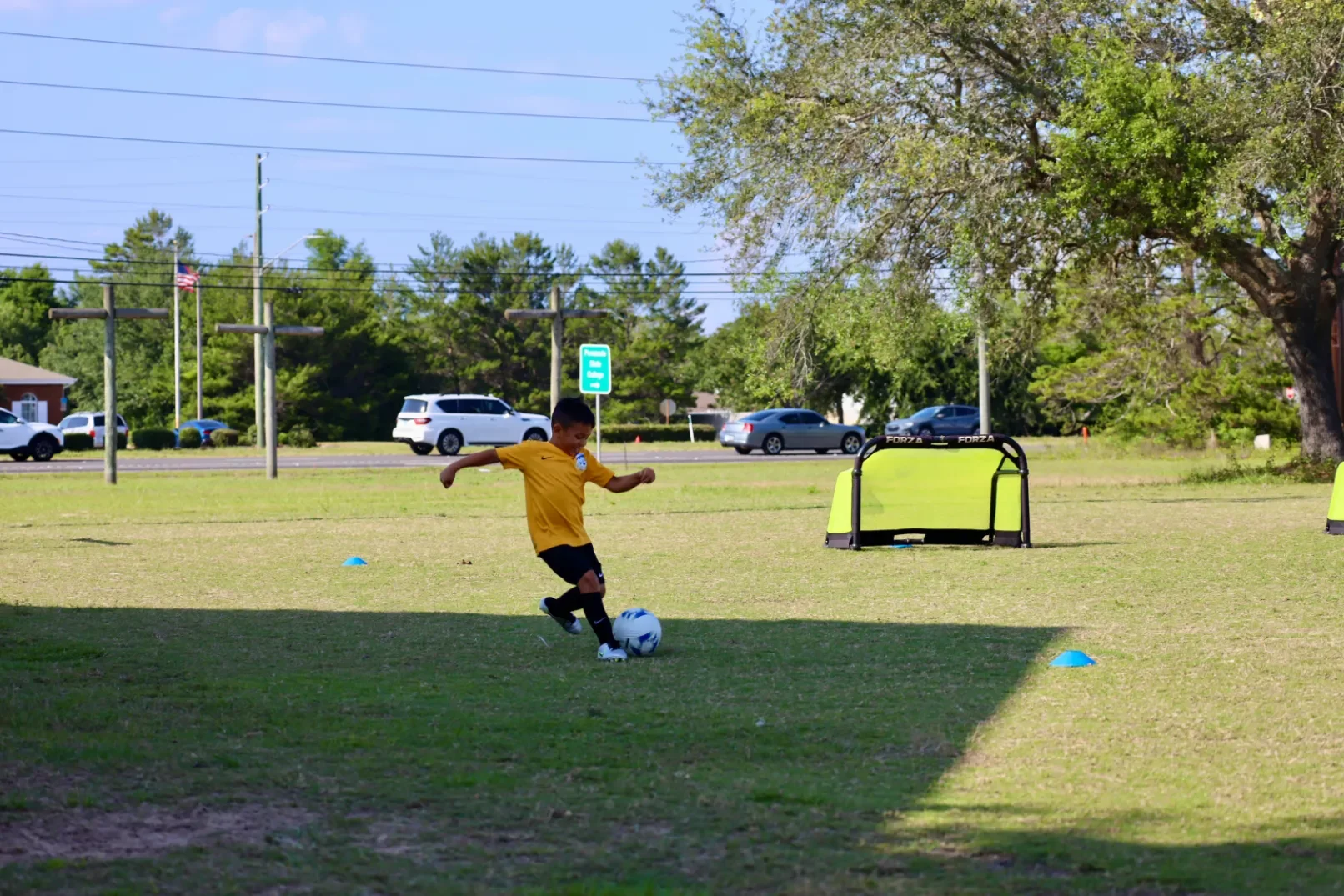A young boy in a yellow shirt and black shorts practicing soccer on a grassy field with small training goals and cones, under a partly shaded sky with trees and parked cars in the background.