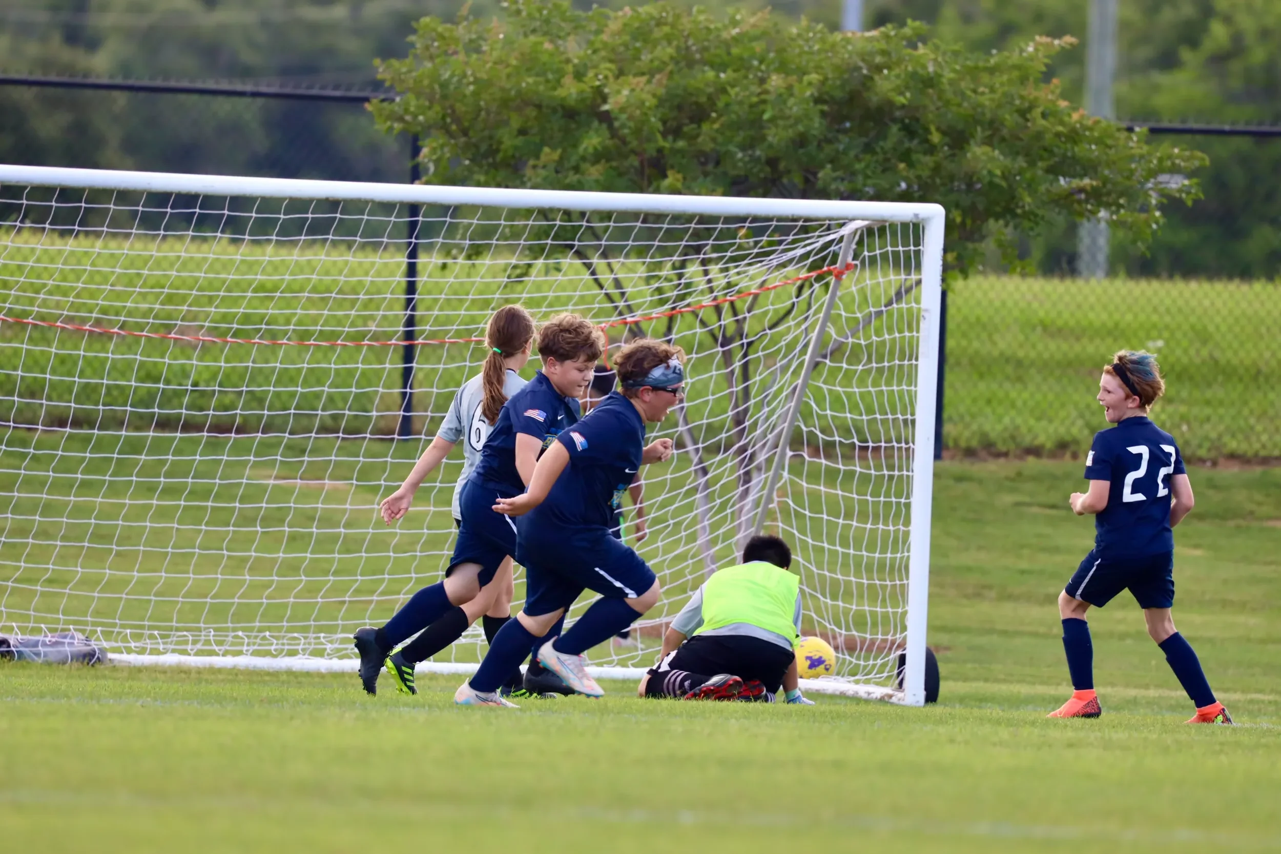 A group of children playing soccer near the goalpost on a grassy field, with some children running and others kneeling near the ground.