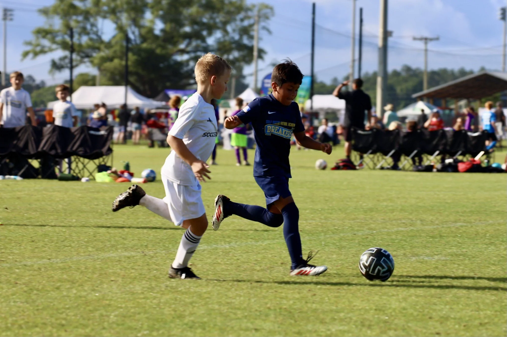 Two young boys playing soccer on a grassy field, chasing after a black and white soccer ball during a sunny day at an outdoor event with spectators and tents in the background.