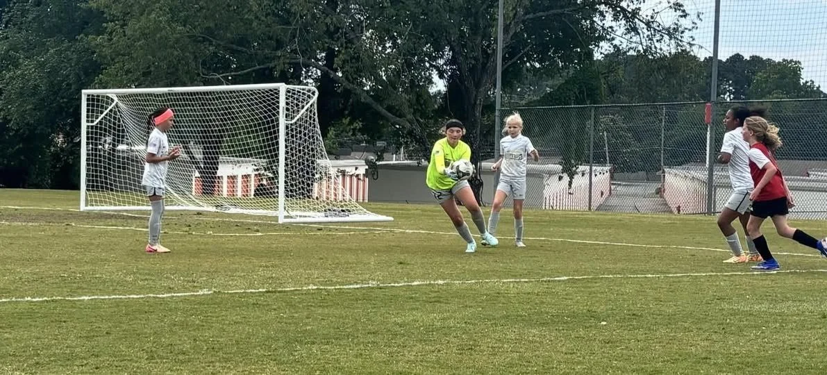 A group of young girls playing soccer on a field, with a goalie holding the ball and other players preparing to kick.