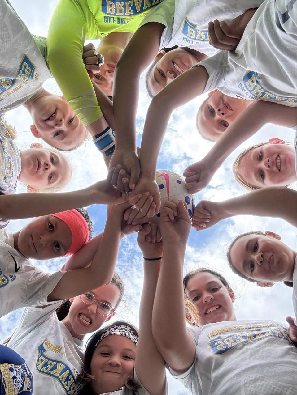 A group of young female soccer players forming a circle with their hands stacked together, holding a soccer ball in the center, taken from below with a background of blue sky and clouds.