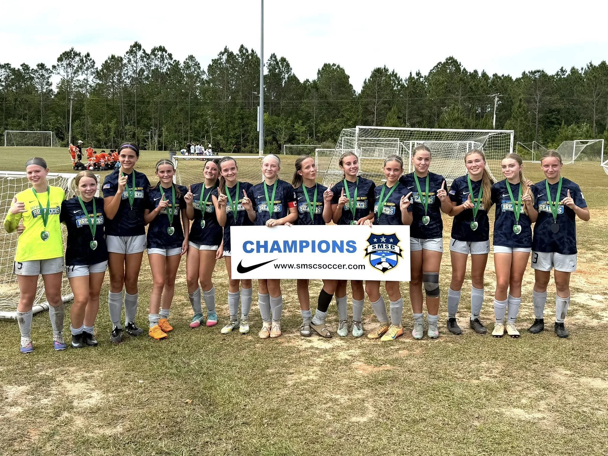 A group of young female soccer players wearing dark blue jerseys and medals, standing together on a grassy field, holding a sign that reads 'Champions' after a soccer game.
