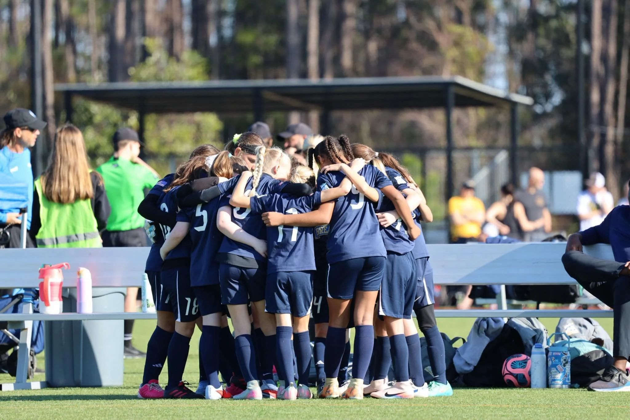 Young female soccer players wearing navy-blue uniforms standing in a circle with arms around each other's shoulders on a field during daytime.