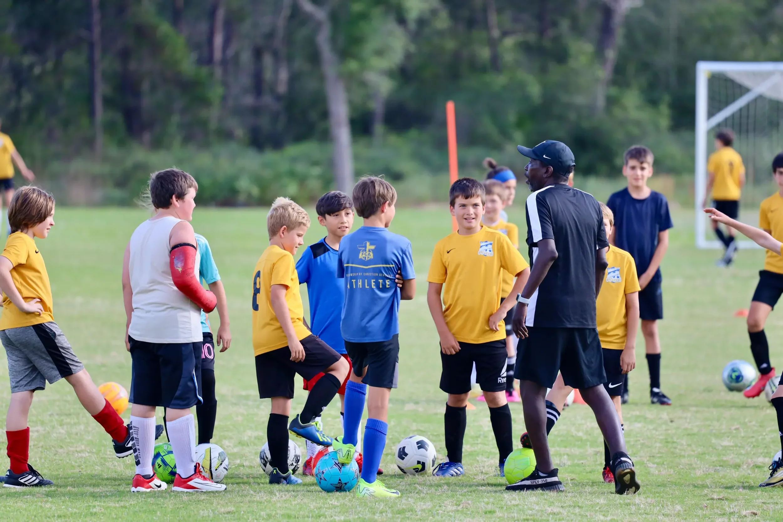 Children in yellow and blue soccer uniforms gathered around a coach during a soccer practice, with soccer balls on the grass field and goalpost in the background.