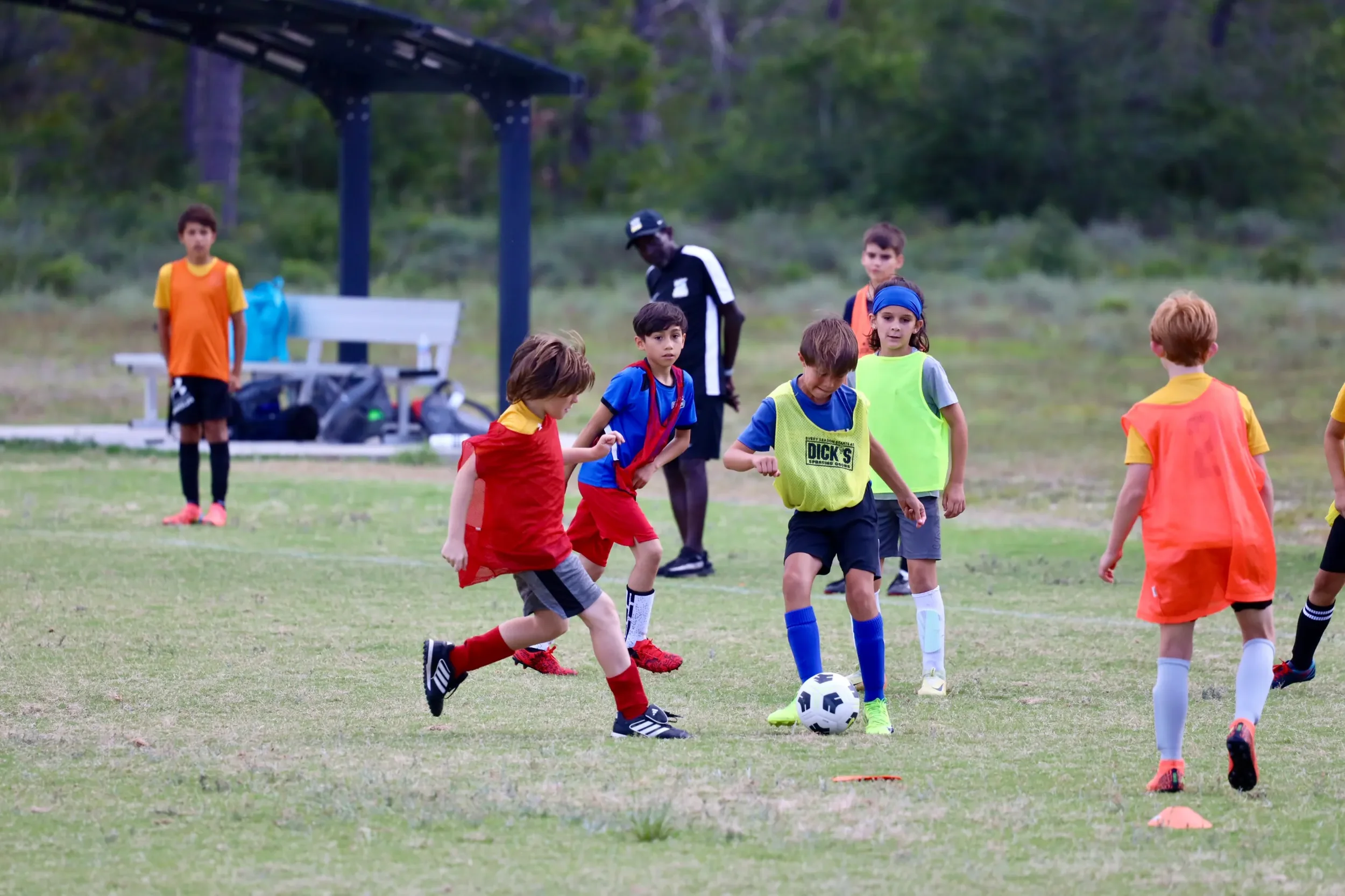 Children playing soccer on a grassy field with a coach in the background.