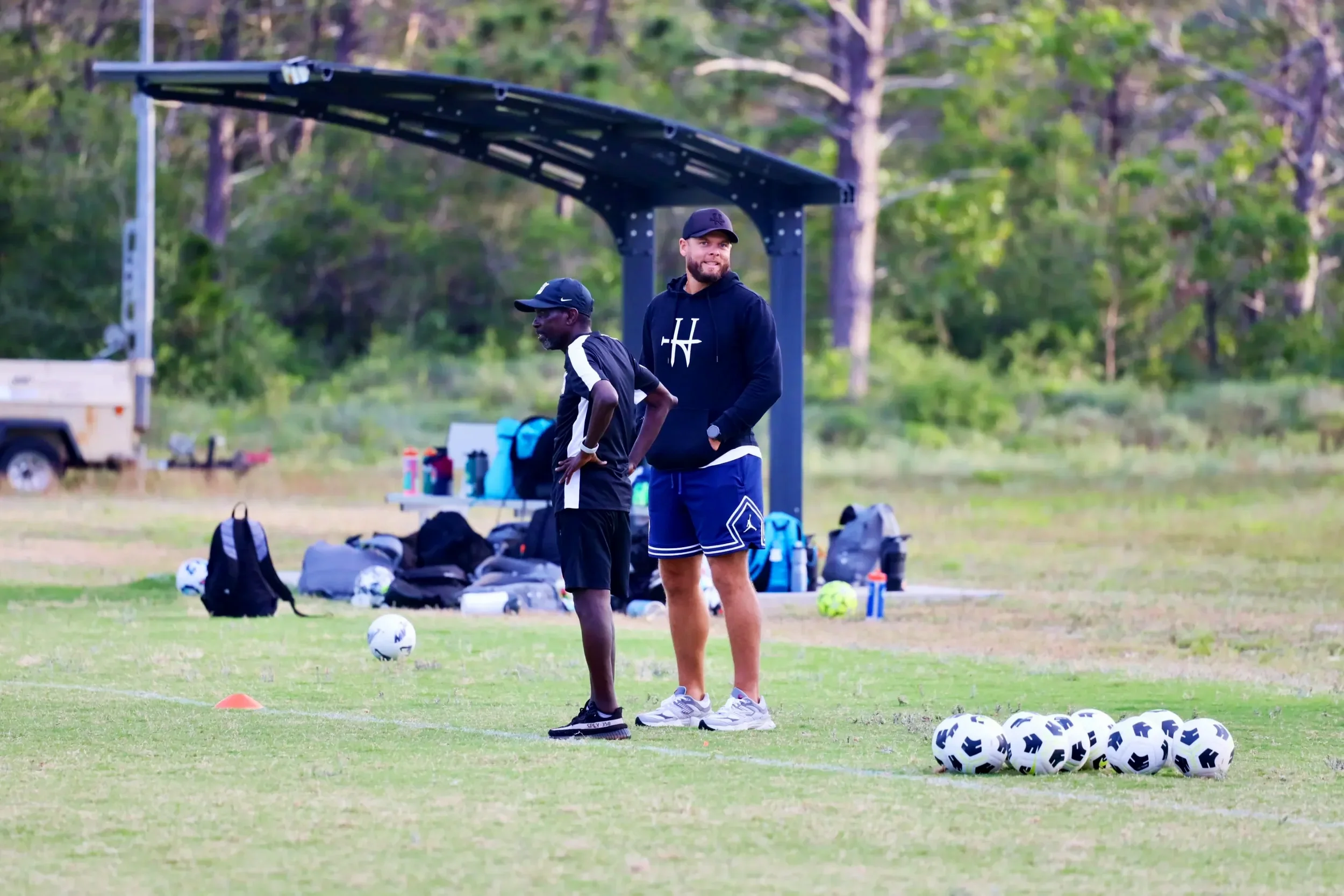 Two men standing on a soccer field, surrounded by soccer balls and equipment, with trees in the background. One man is wearing a black hoodie and shorts, and the other is in a black shirt and shorts.